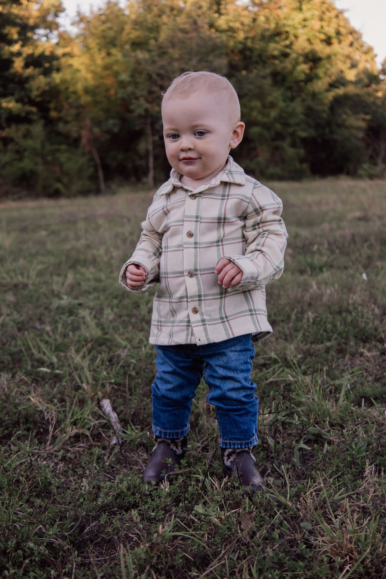 A small child wearing a plaid button-up shirt and jeans stands in a grassy field during golden hour.