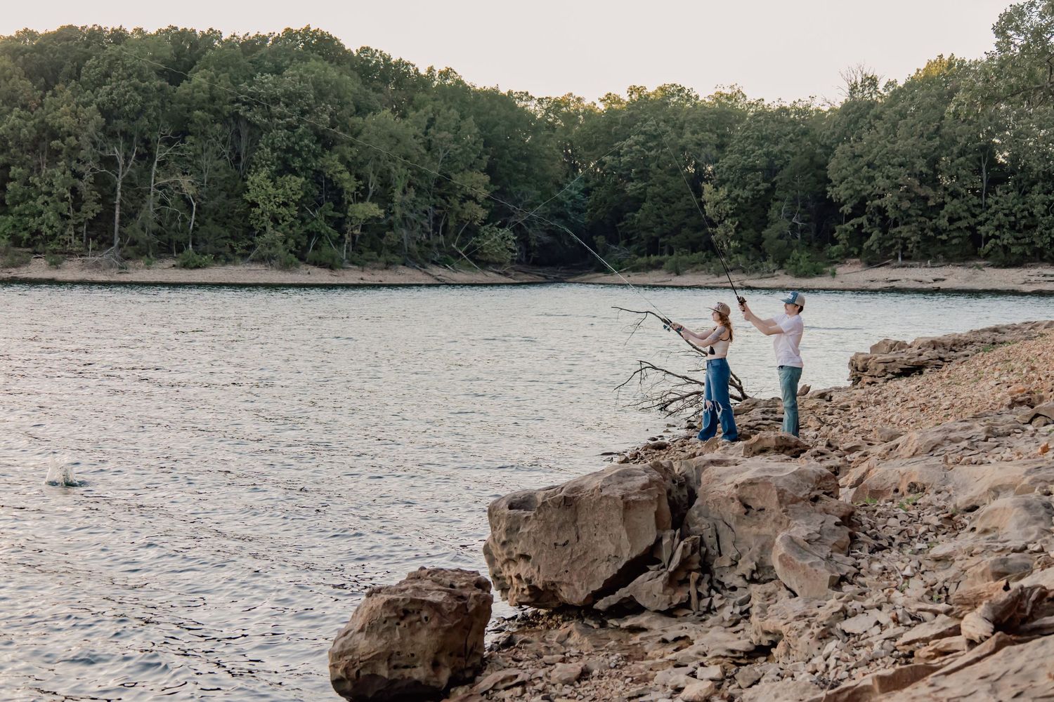 People fishing from rocky shoreline at lake during sunset with trees in background.