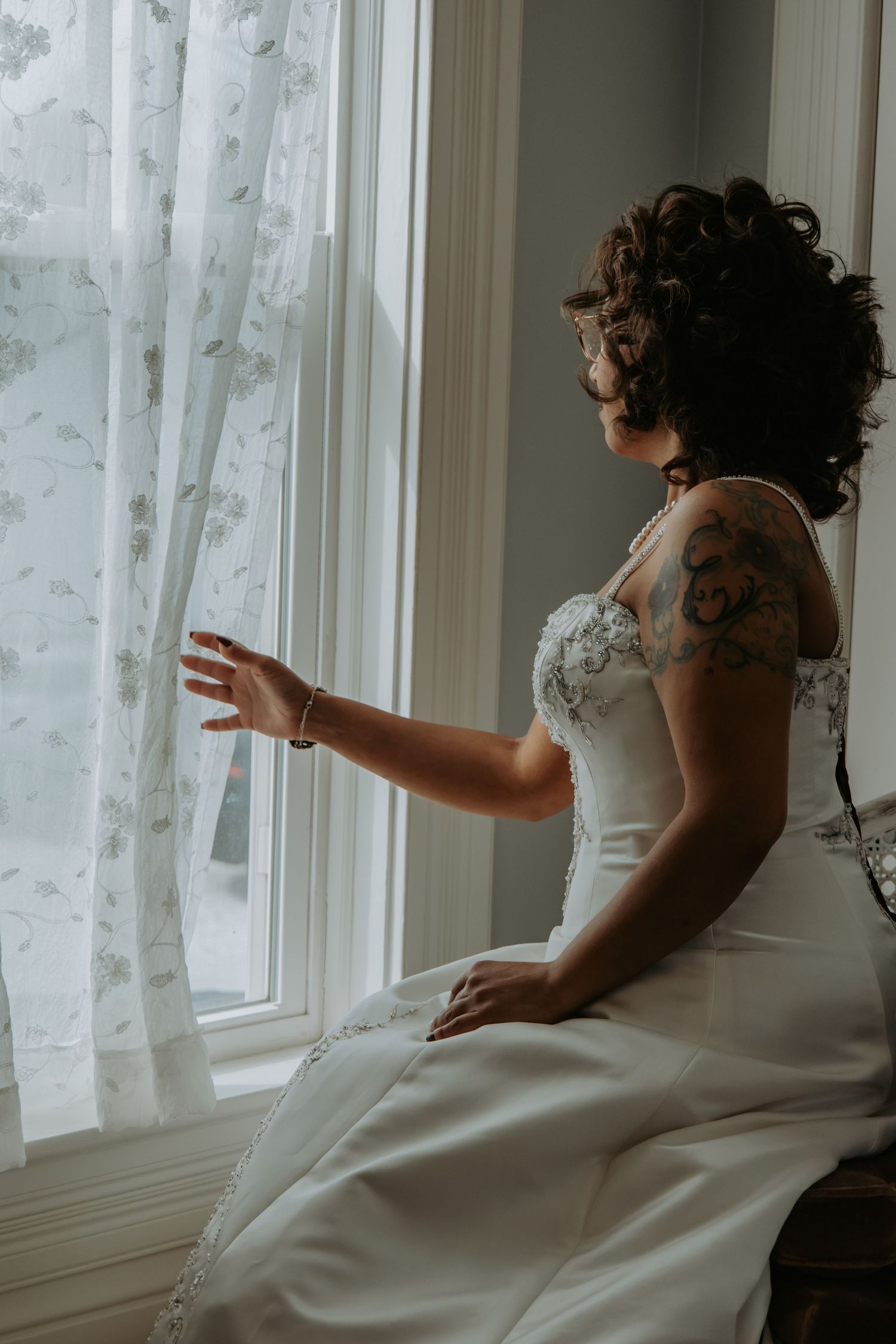 Bride in white gown with tattoos gazing through window with sheer curtains in natural light.