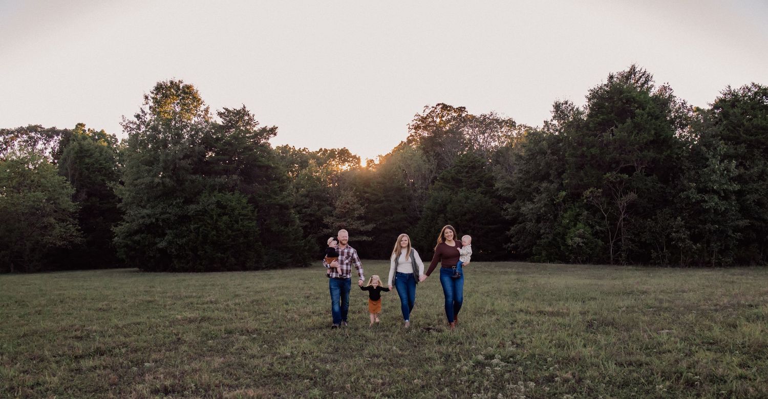 A panoramic view of a grassy field at sunset with trees in the background and figures silhouetted against the sky.