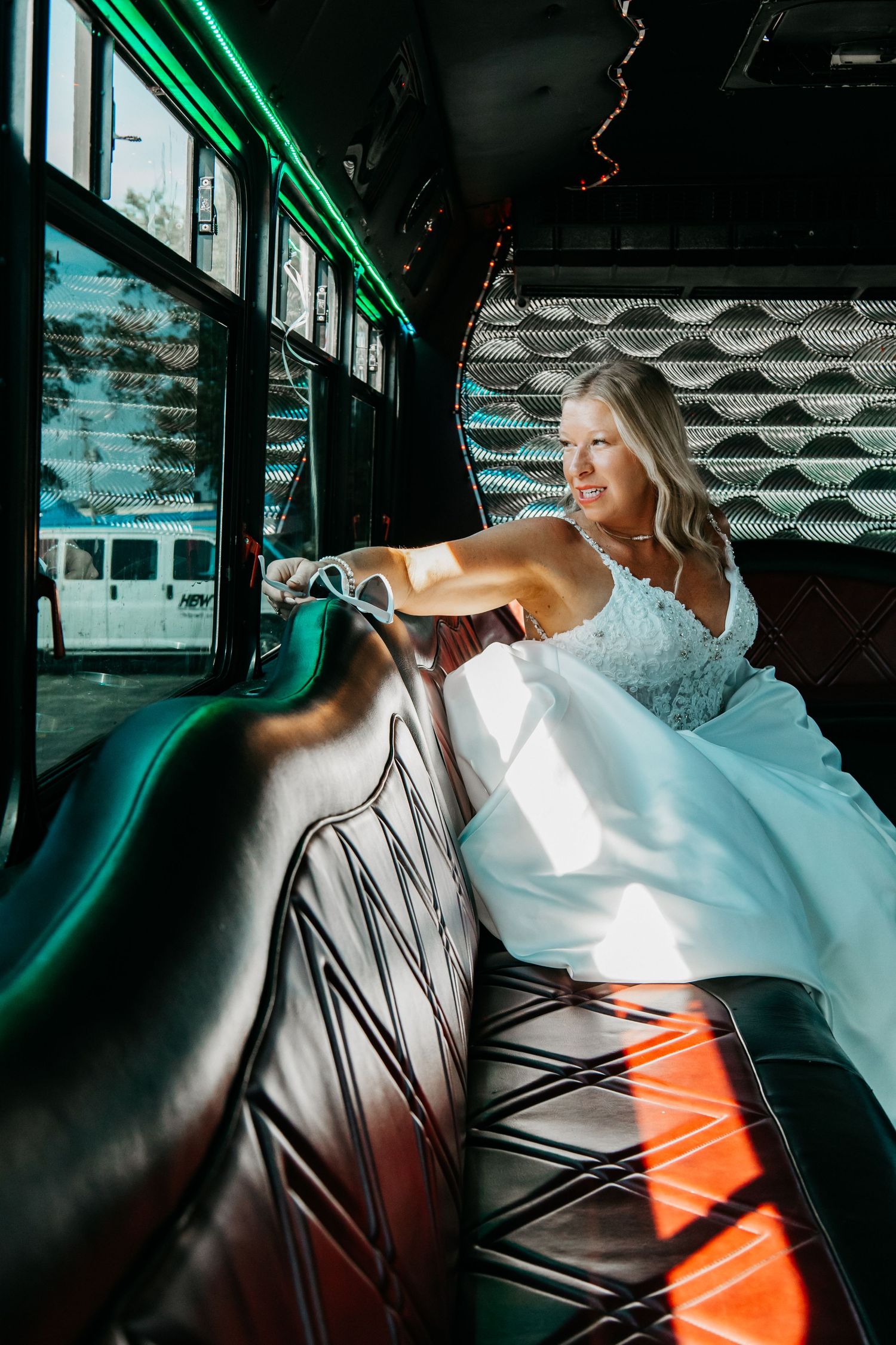 A bride in a white dress reclines on dark leather seating with dramatic green lighting in what appears to be a party bus.