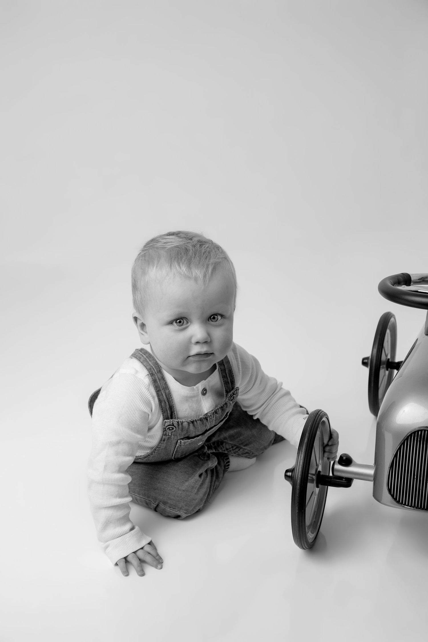 A small child in denim overalls and white shirt crawls beside a vintage toy pedal car in black and white photography.