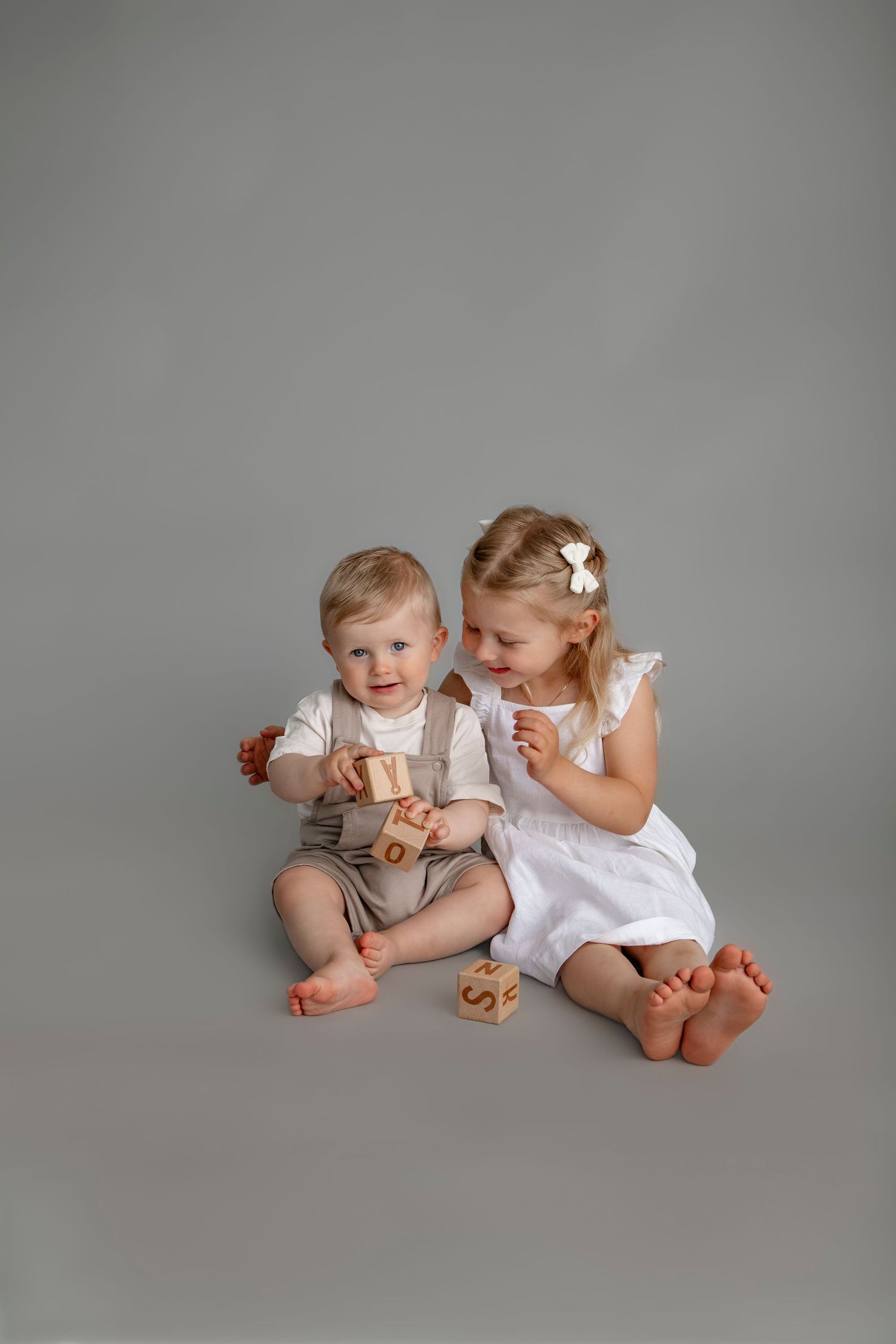 Two young children in white outfits sit and laugh together on a light gray backdrop during a studio photo session.