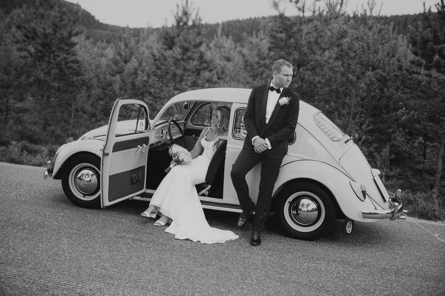 A classic Volkswagen Beetle parked on a mountain road serves as backdrop for a romantic wedding photo in black and white.