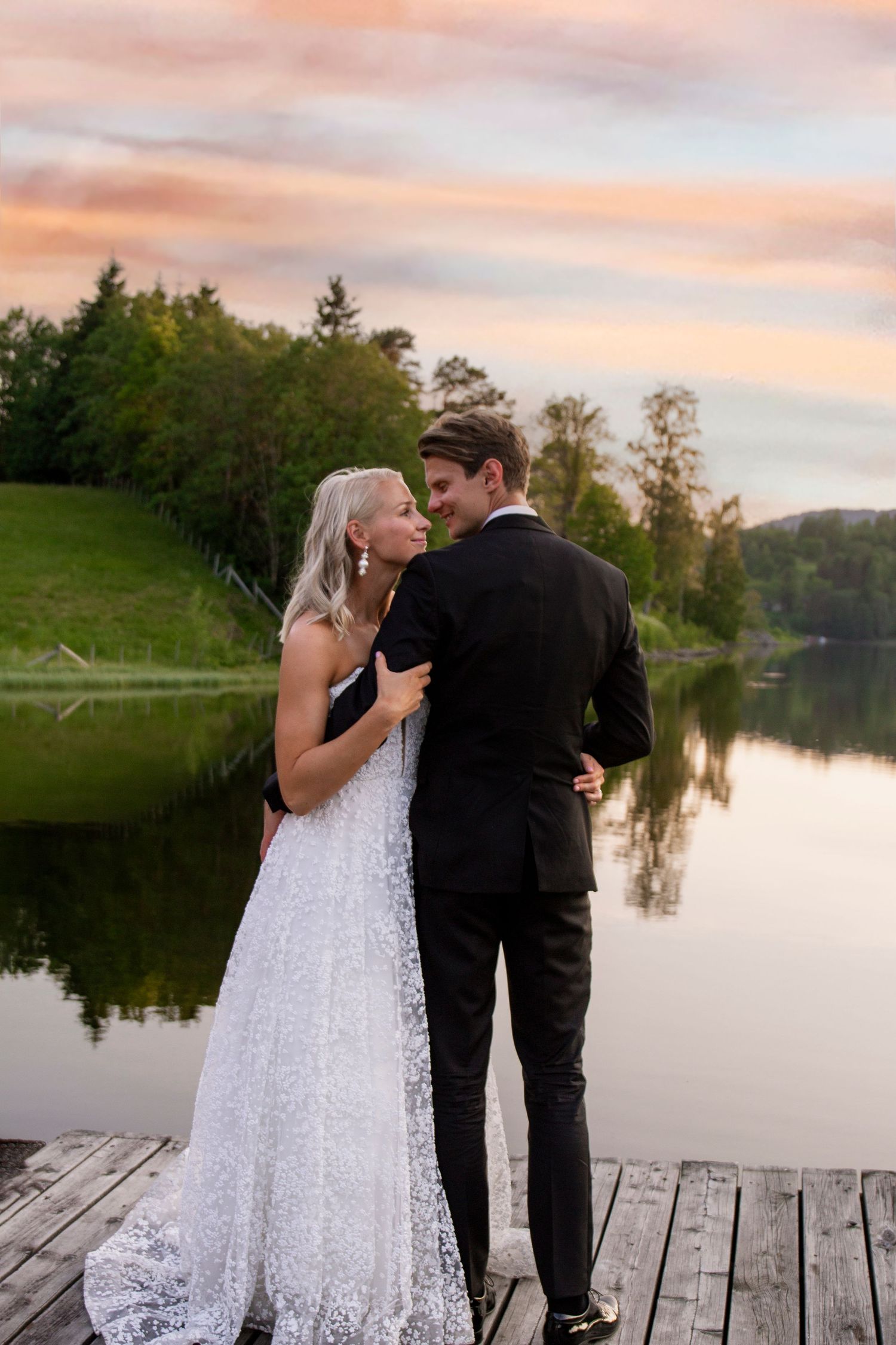 Newlywed couple embraces on wooden dock by serene lake at sunset with forest backdrop.
