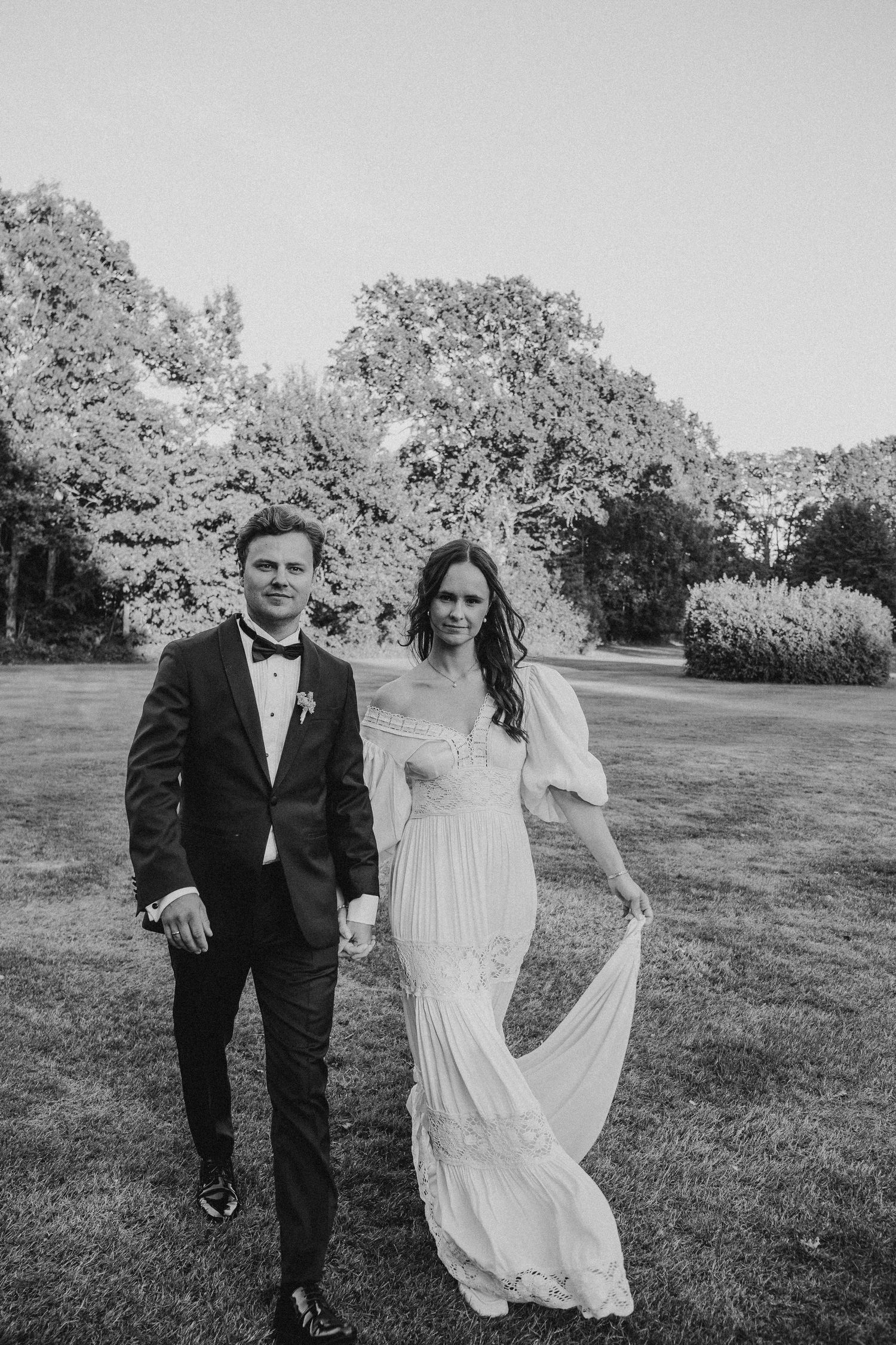 A black and white wedding photo of a couple standing on a lawn with trees in the background.