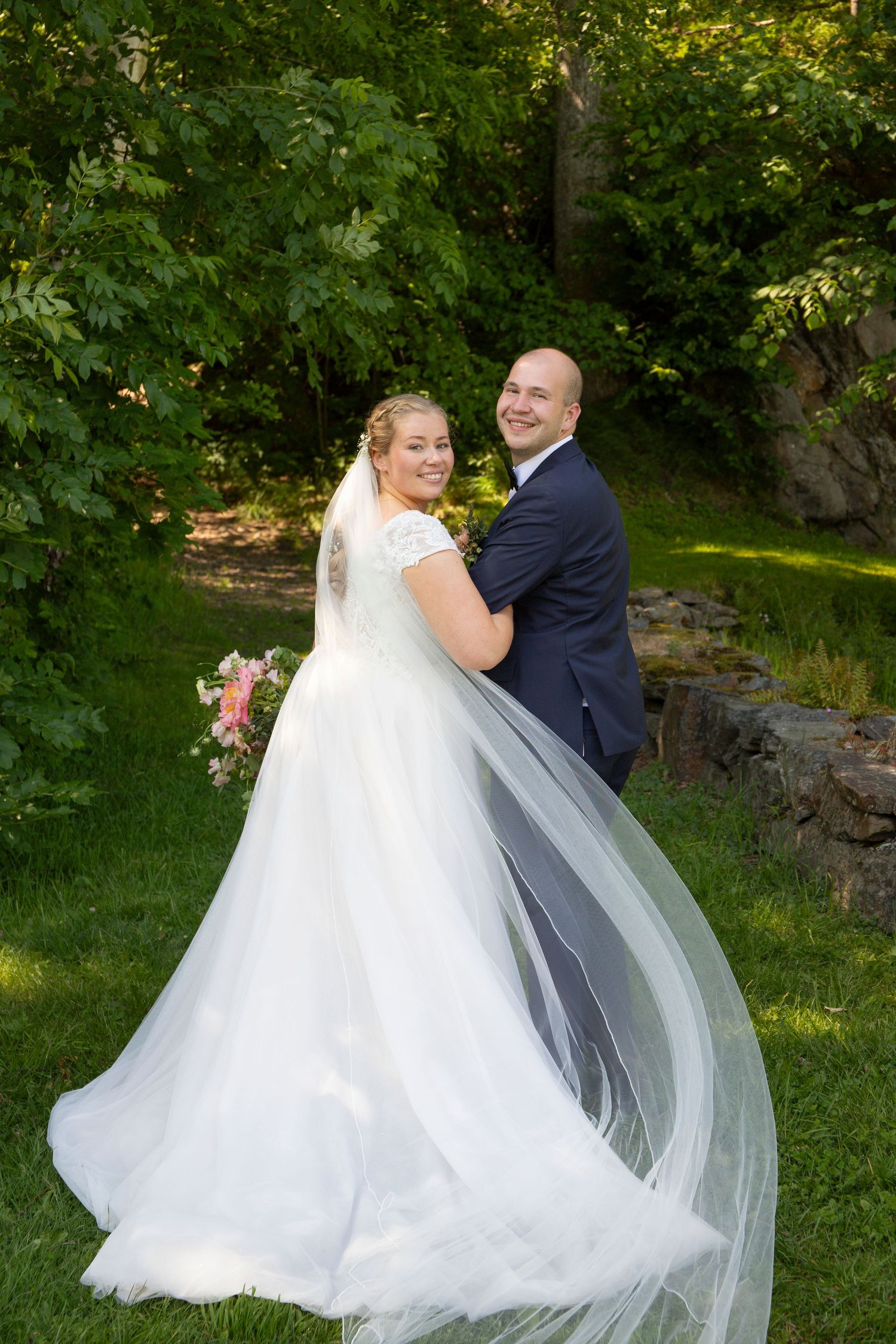 A couple poses in a garden setting on their wedding day, with a flowing white gown and navy suit against lush greenery.