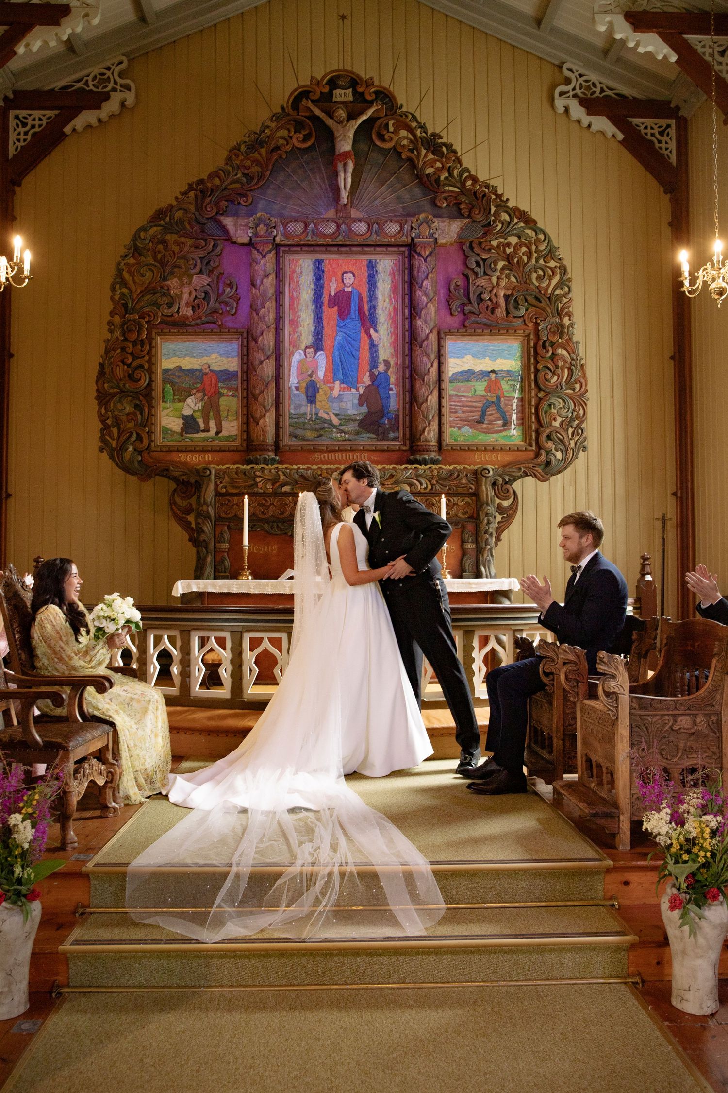 A wedding ceremony takes place in a beautiful church with ornate religious artwork and stained glass windows illuminating the altar.
