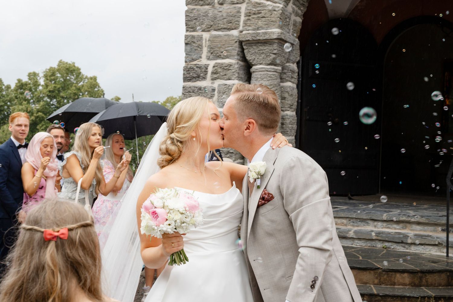 Newlywed couple celebrates with bubbles outside a stone church while wedding guests look on.