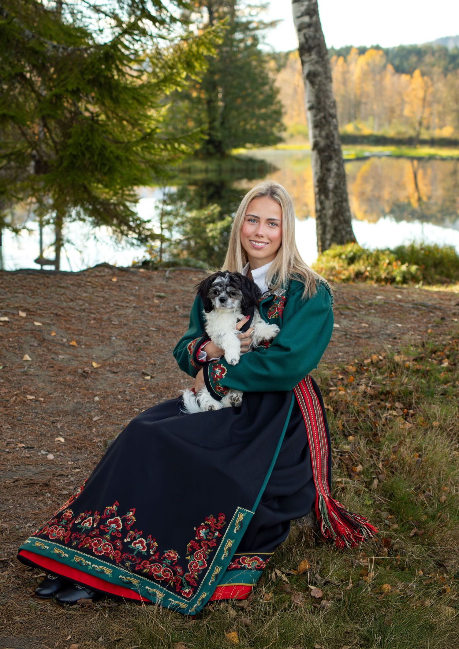 A person in a green sweater sits with a black and white cat on a forest floor covered in pine needles by a lake.