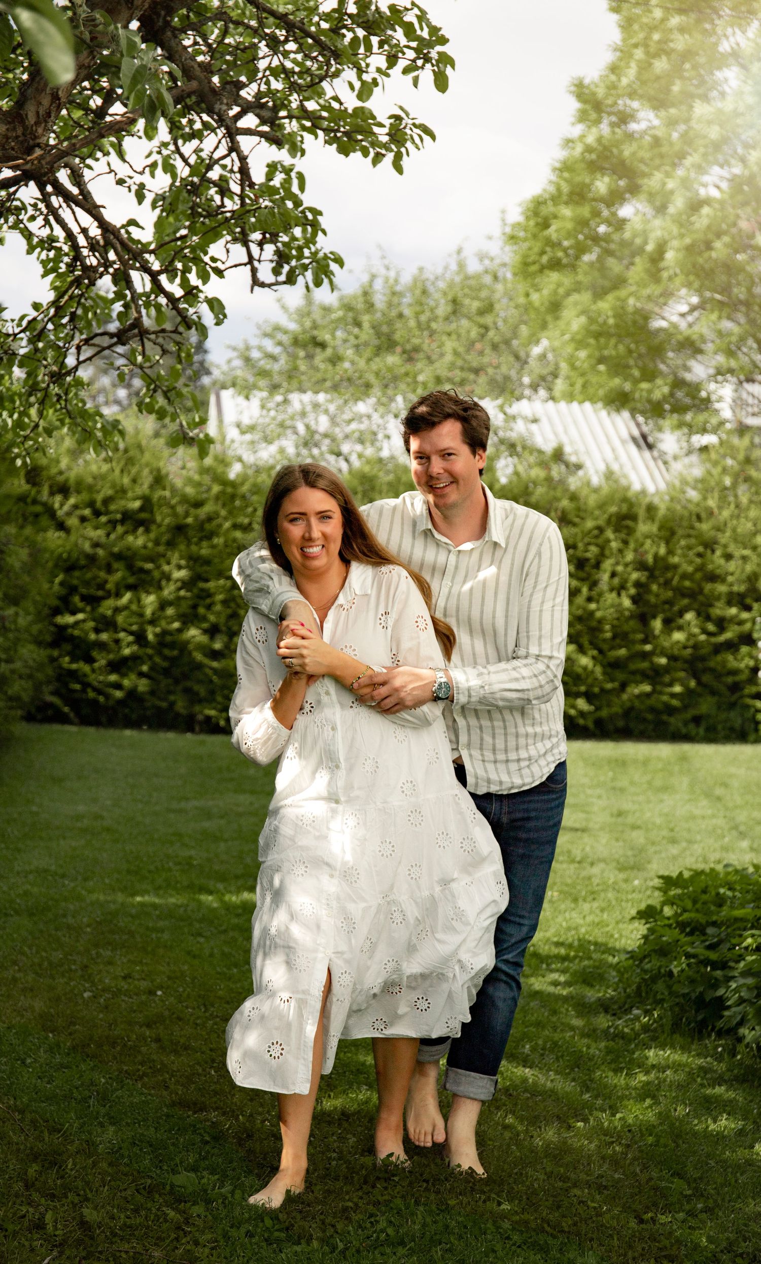 A playful couple poses together on a green lawn under trees on a sunny summer day.