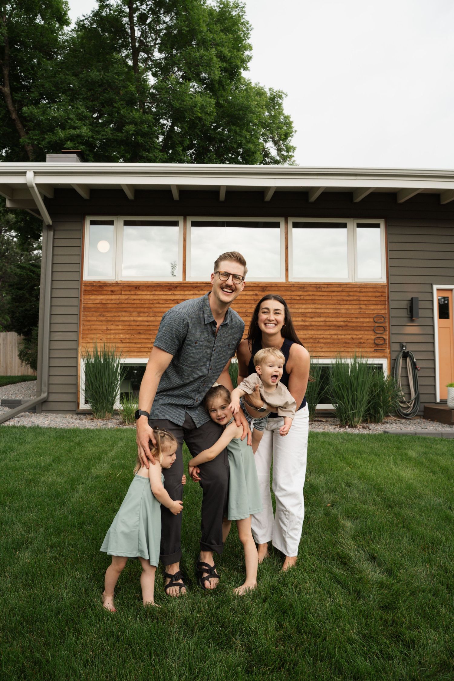 Happy family of five posing together on green lawn in front of modern mid-century home on cloudy day.
