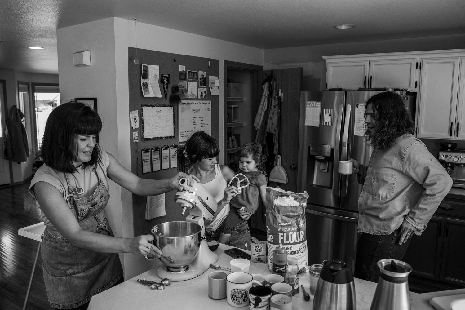 Family baking together in kitchen, black and white photo of women and children mixing ingredients at counter.