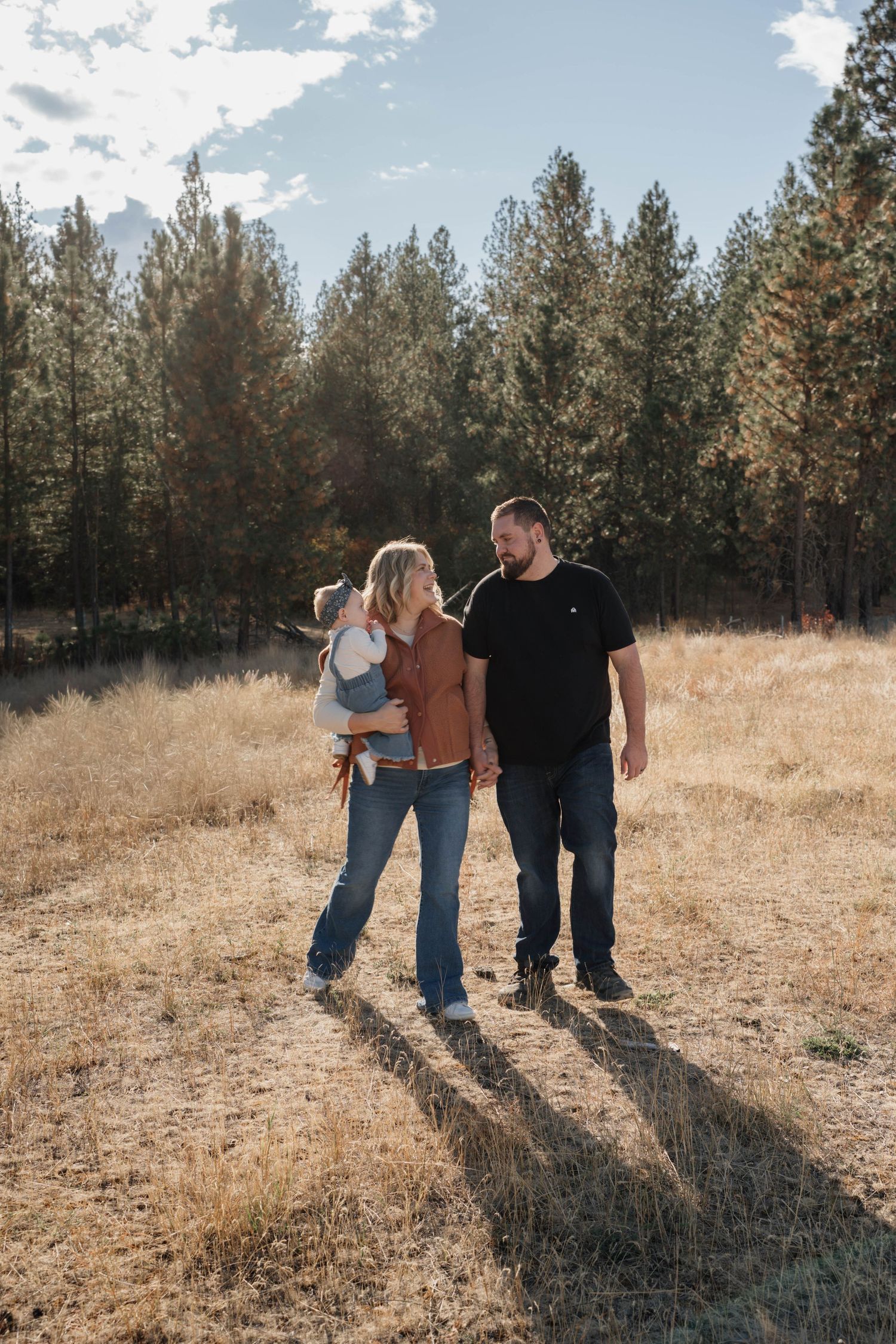 Three people walk together through a golden field with evergreen trees in the background on a sunny afternoon.