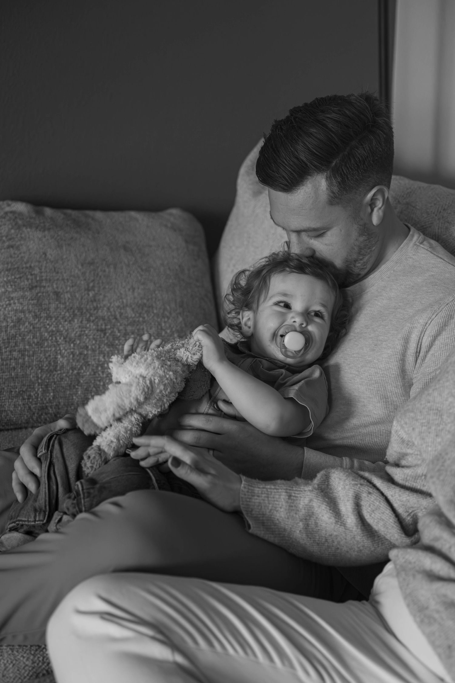 Black and white photo of a father holding his toddler with a pacifier and teddy bear on a cozy sofa.