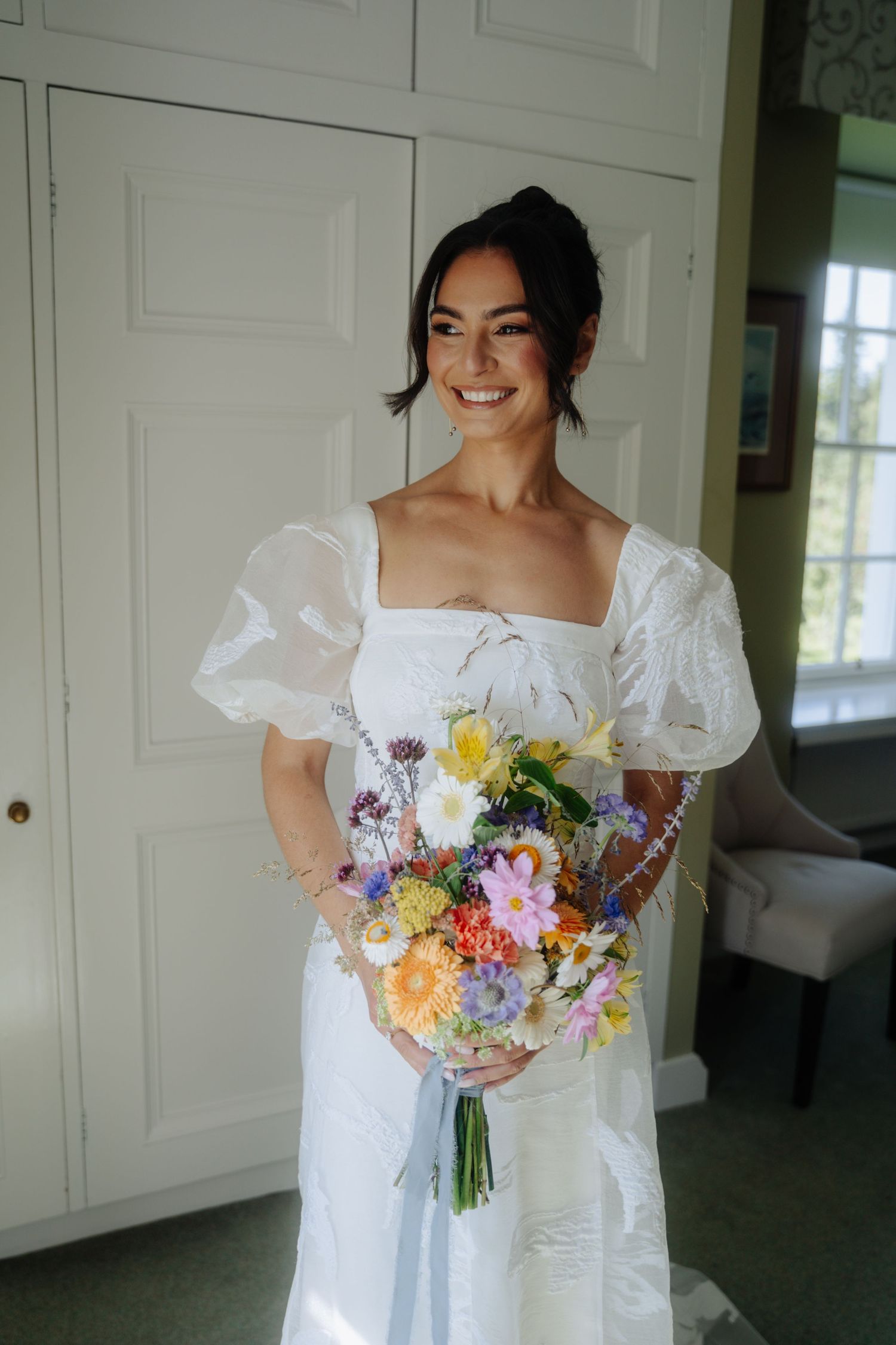 A bride in a white dress with puff sleeves holds a vibrant summer bouquet of mixed flowers.