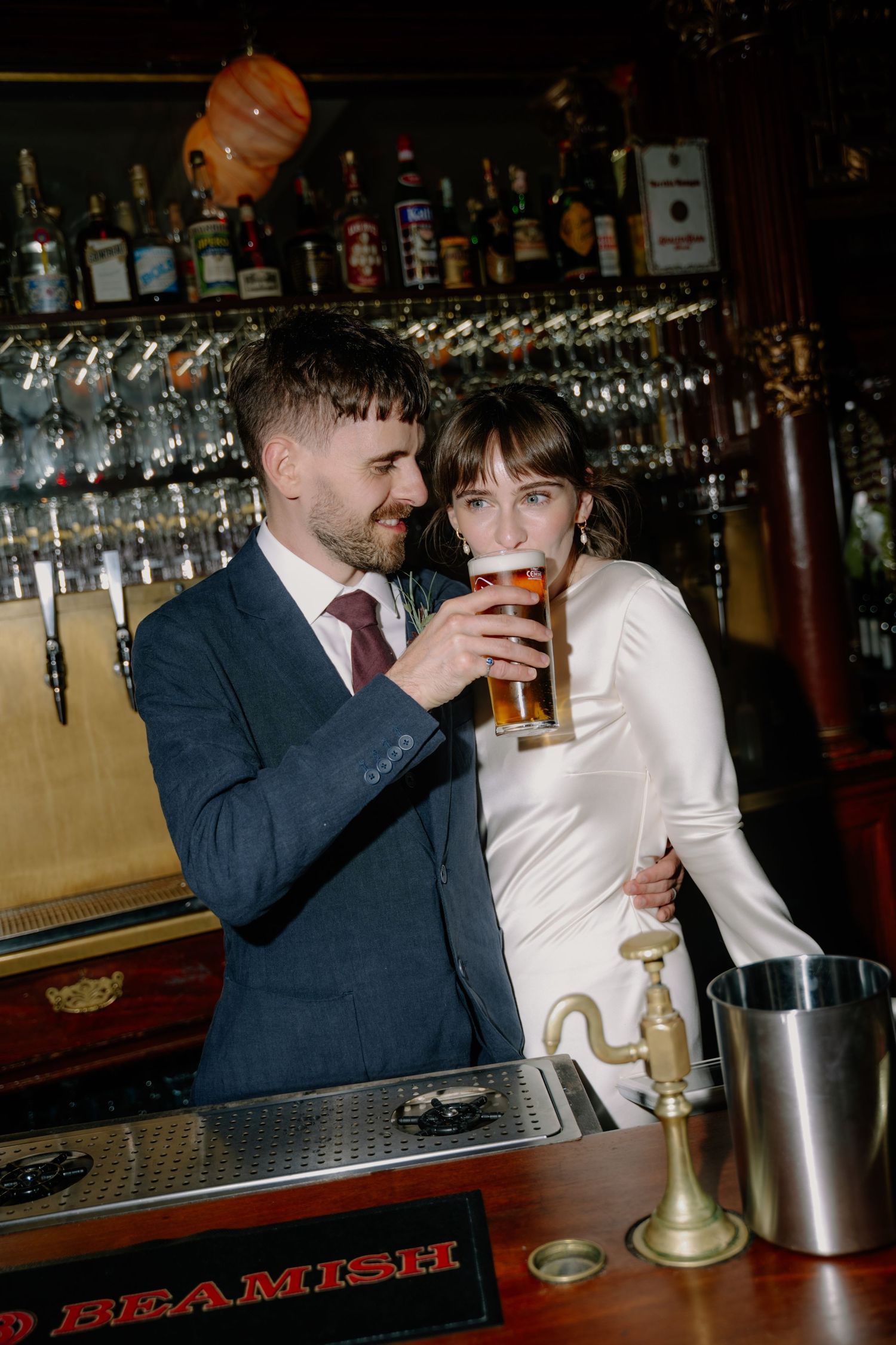 Bride and groom share a beer at a pub bar on their wedding day, smiling together in wedding attire.