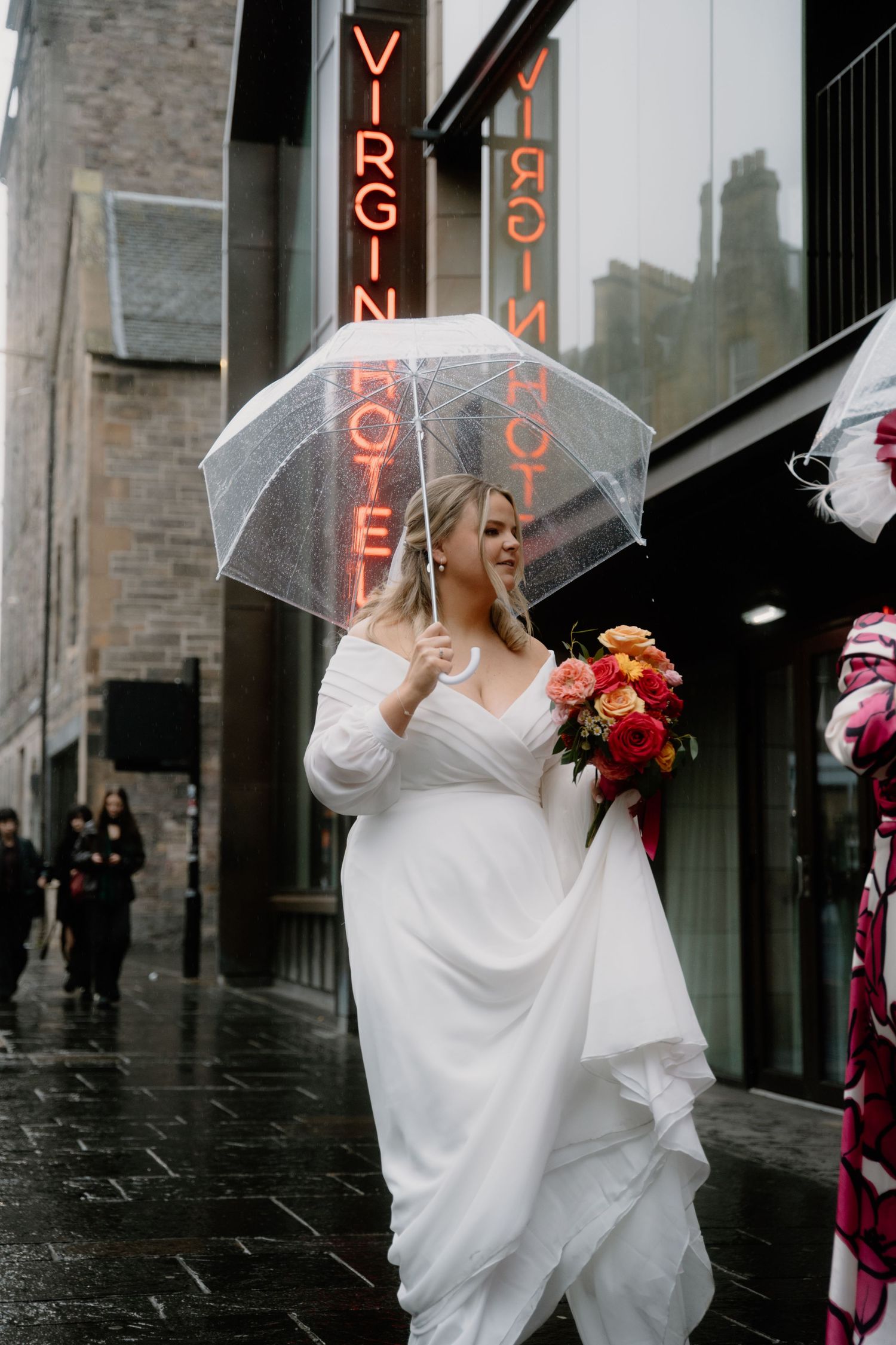 A person in a white dress holds clear umbrella and bouquet outside Virgin Hotel Edinburgh on rainy day.