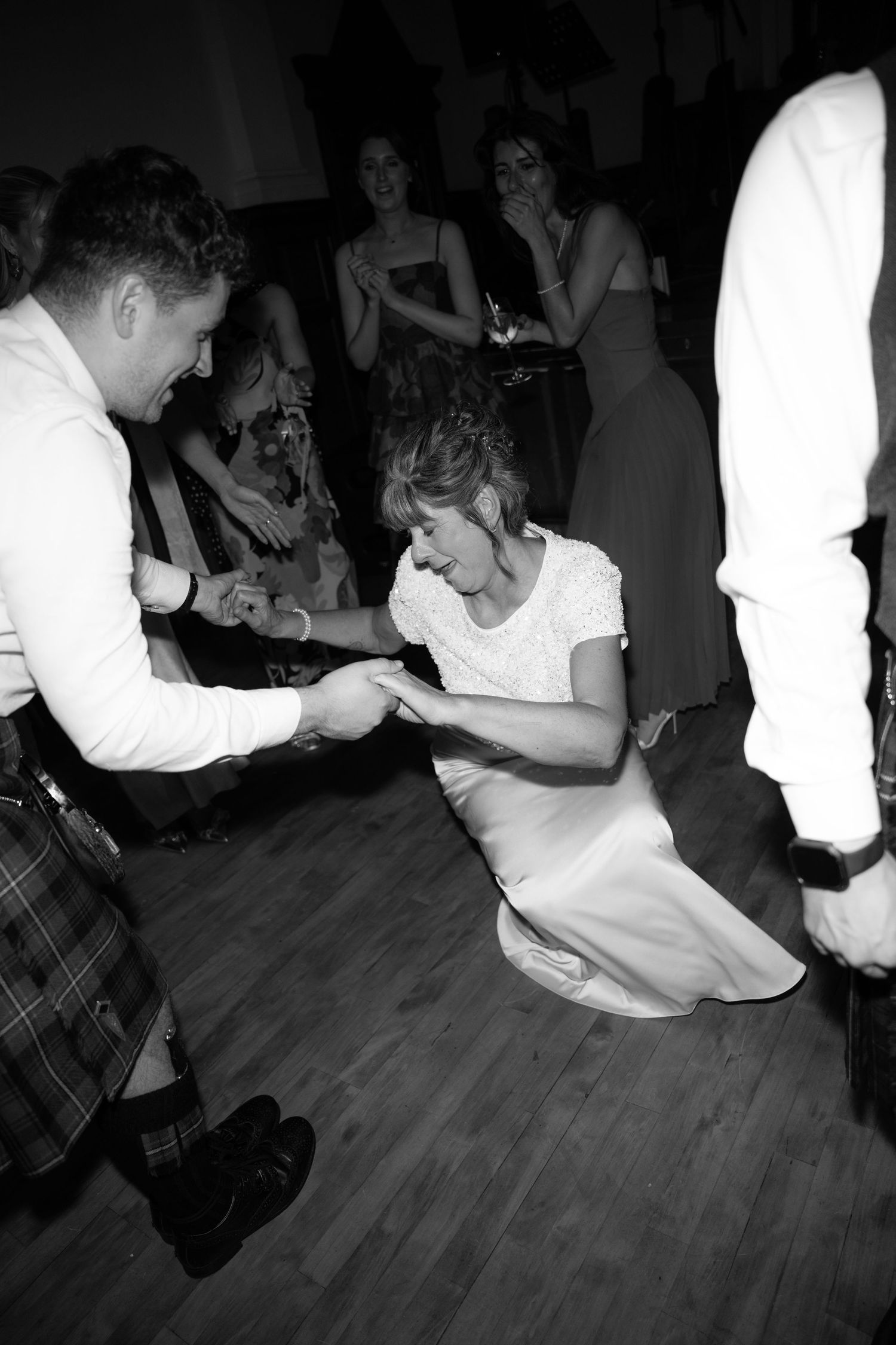 Black and white photo of a bride crouching on dance floor holding hands with a kilted groom at a Scottish wedding reception.