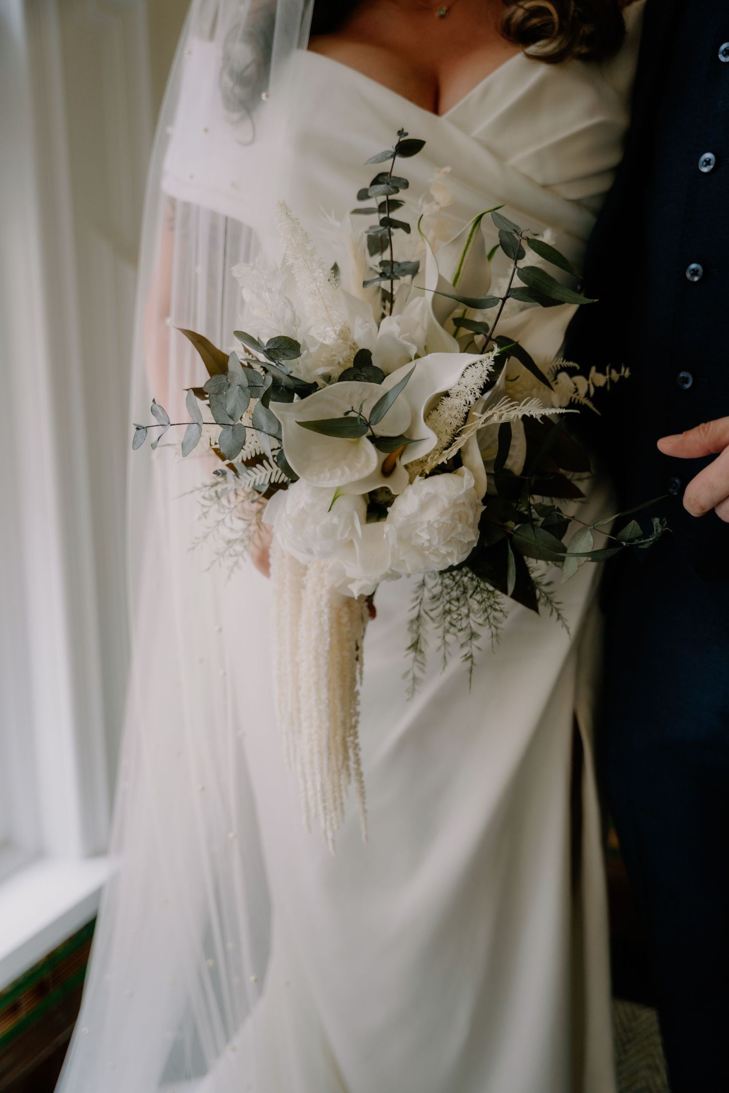Bride holding elegant white wedding bouquet with calla lilies, eucalyptus, and pampas grass, wearing a flowing gown and veil.