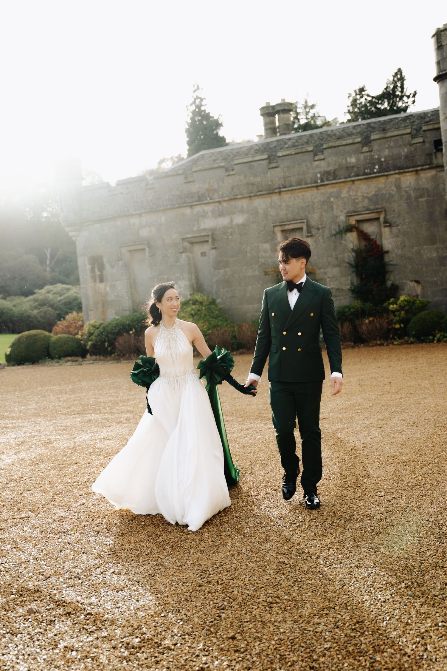 Bride in white gown and groom in black tuxedo walk hand in hand on gravel path outside stone manor house.