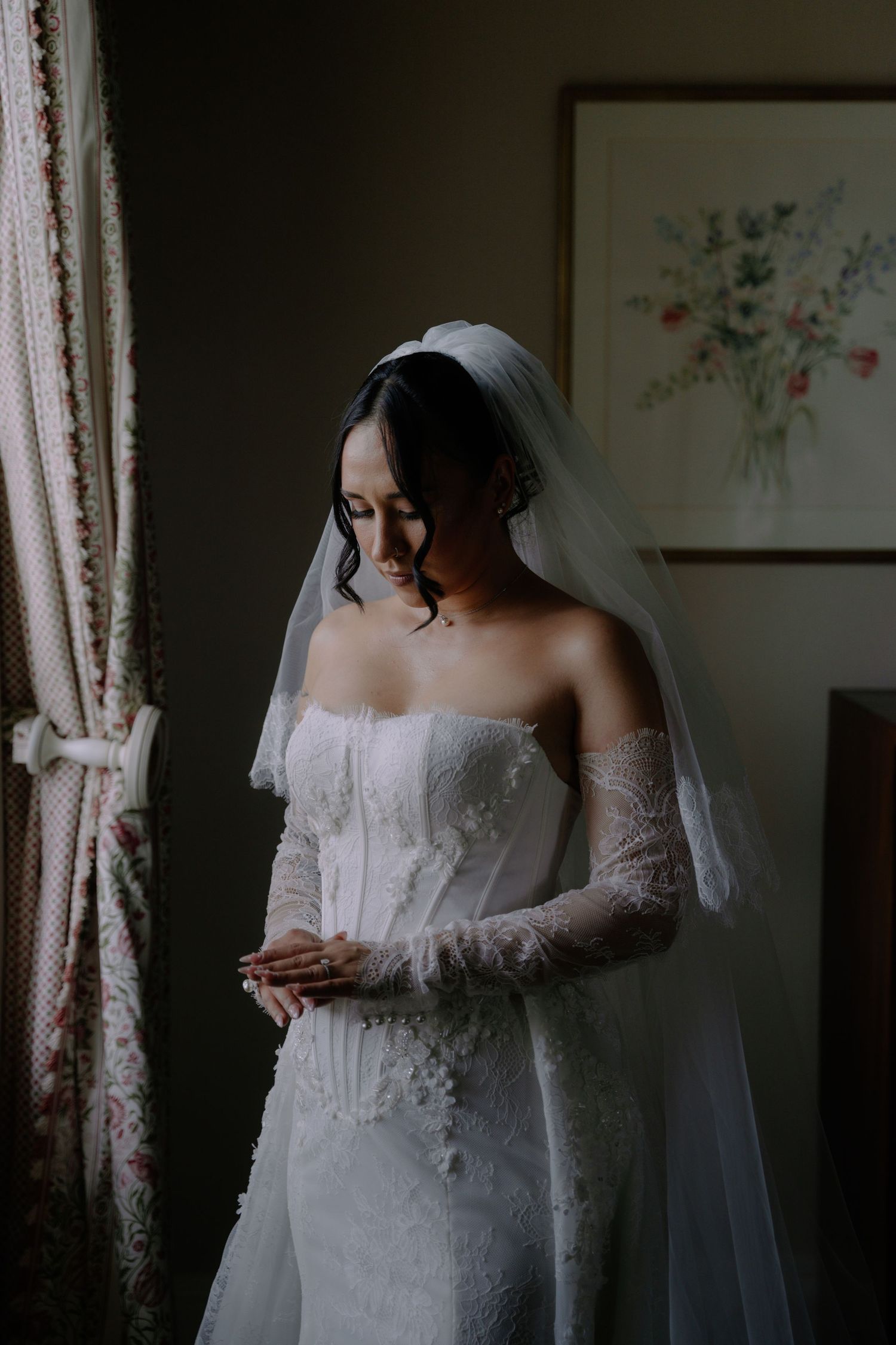Bride in strapless lace wedding gown with sheer sleeves and veil, standing by window in soft natural light.