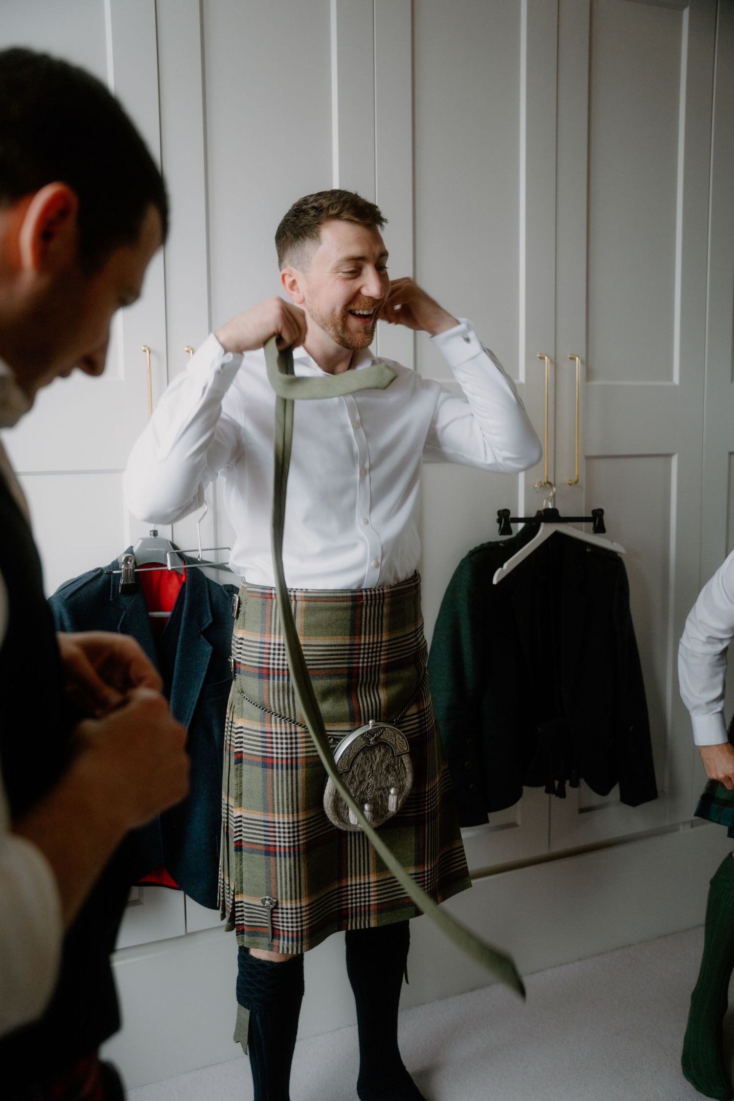 Person wearing a white dress shirt and traditional plaid kilt getting dressed in formal Scottish attire.