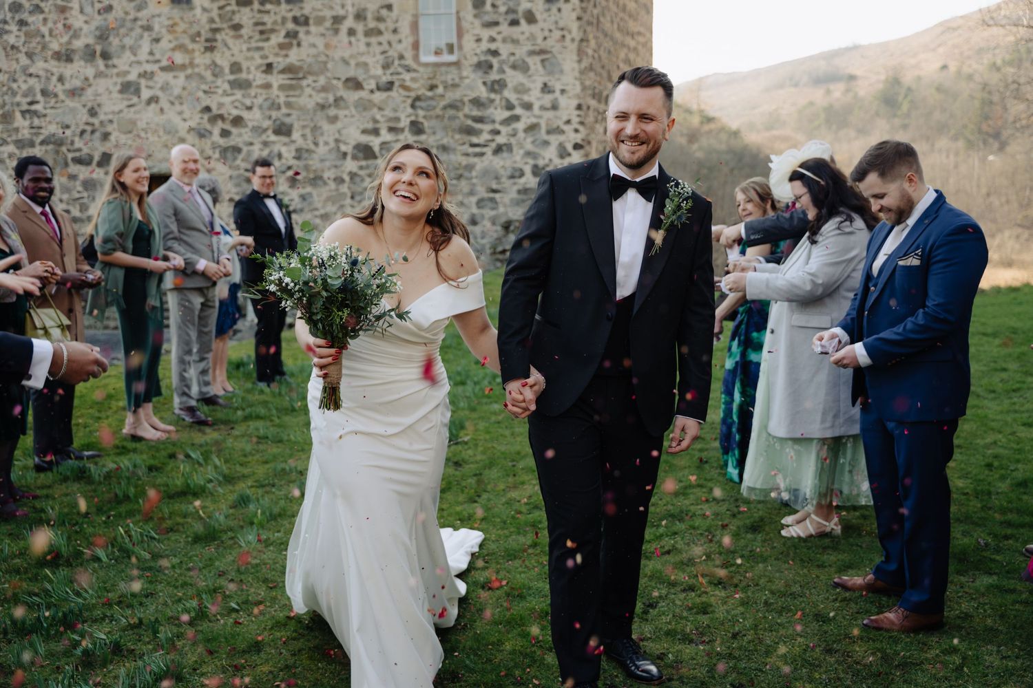 Wedding guests shower newlyweds with confetti during outdoor ceremony at rustic stone venue.