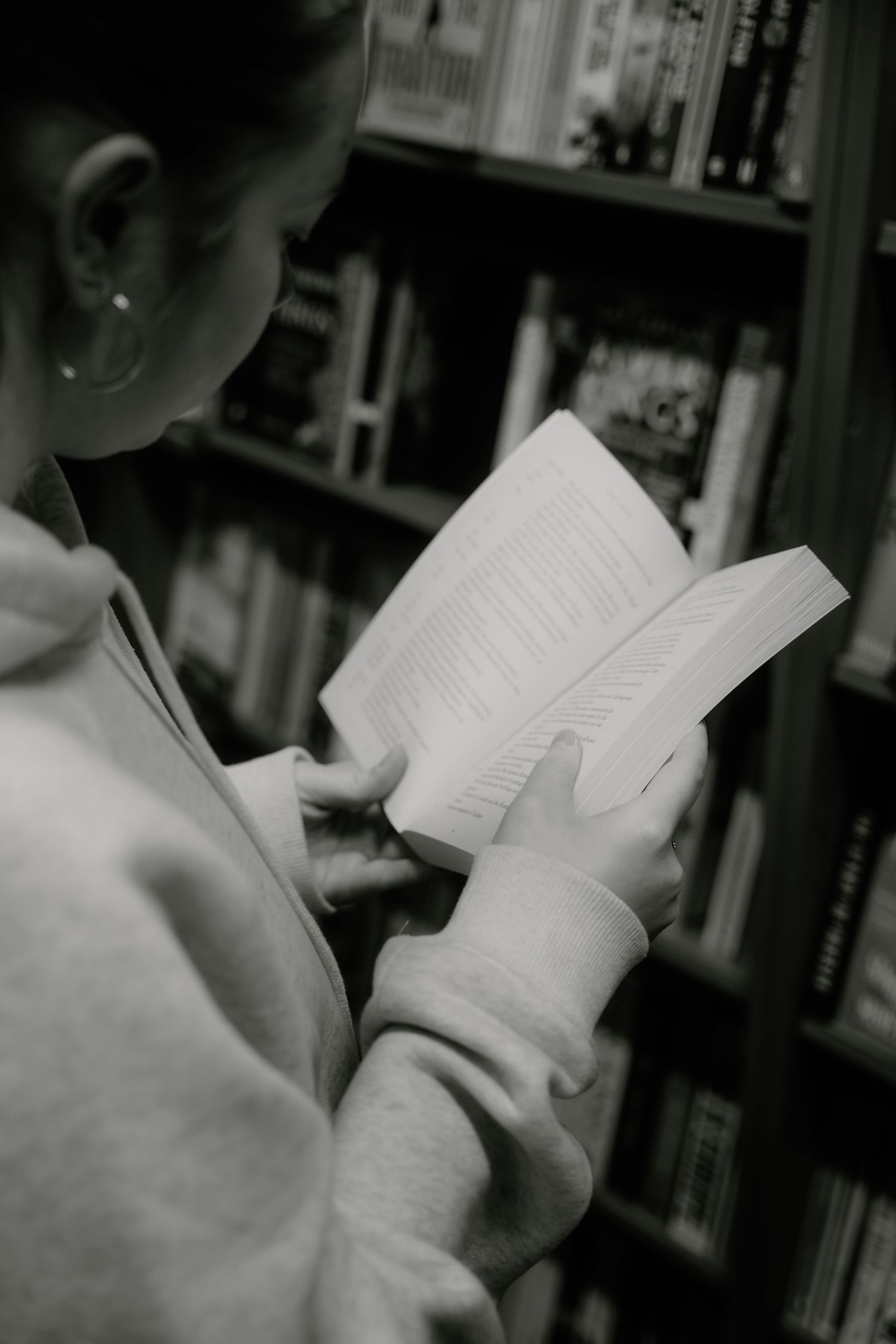 Black and white photo of someone reading a book in front of a bookshelf.