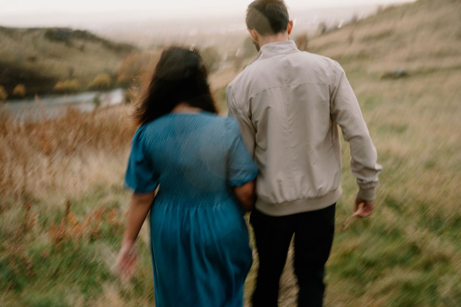 A couple in casual clothing walks together through a golden grass field on a cloudy autumn day.