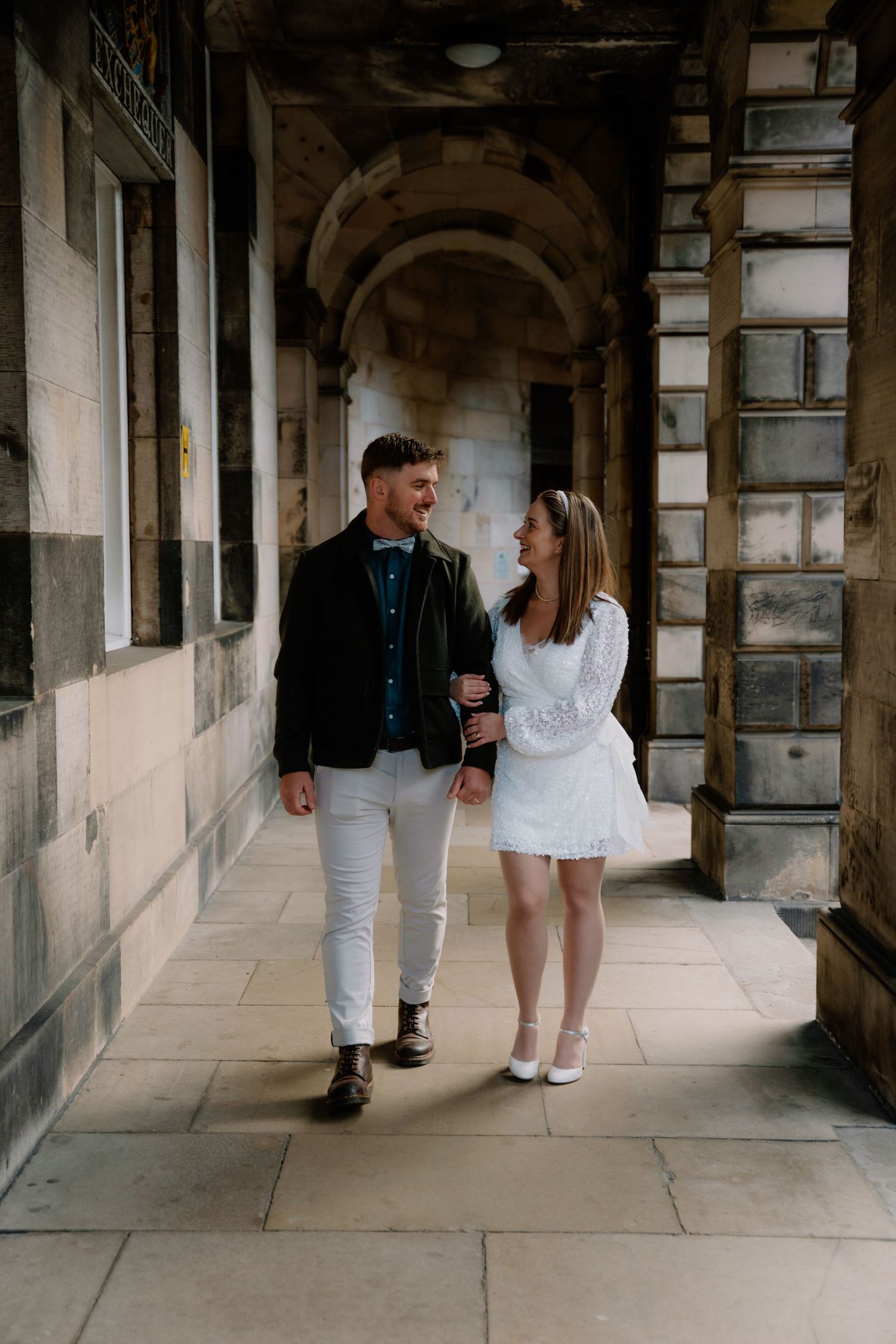 A couple walks through a historic stone corridor with arched windows and natural lighting casting shadows on the floor.