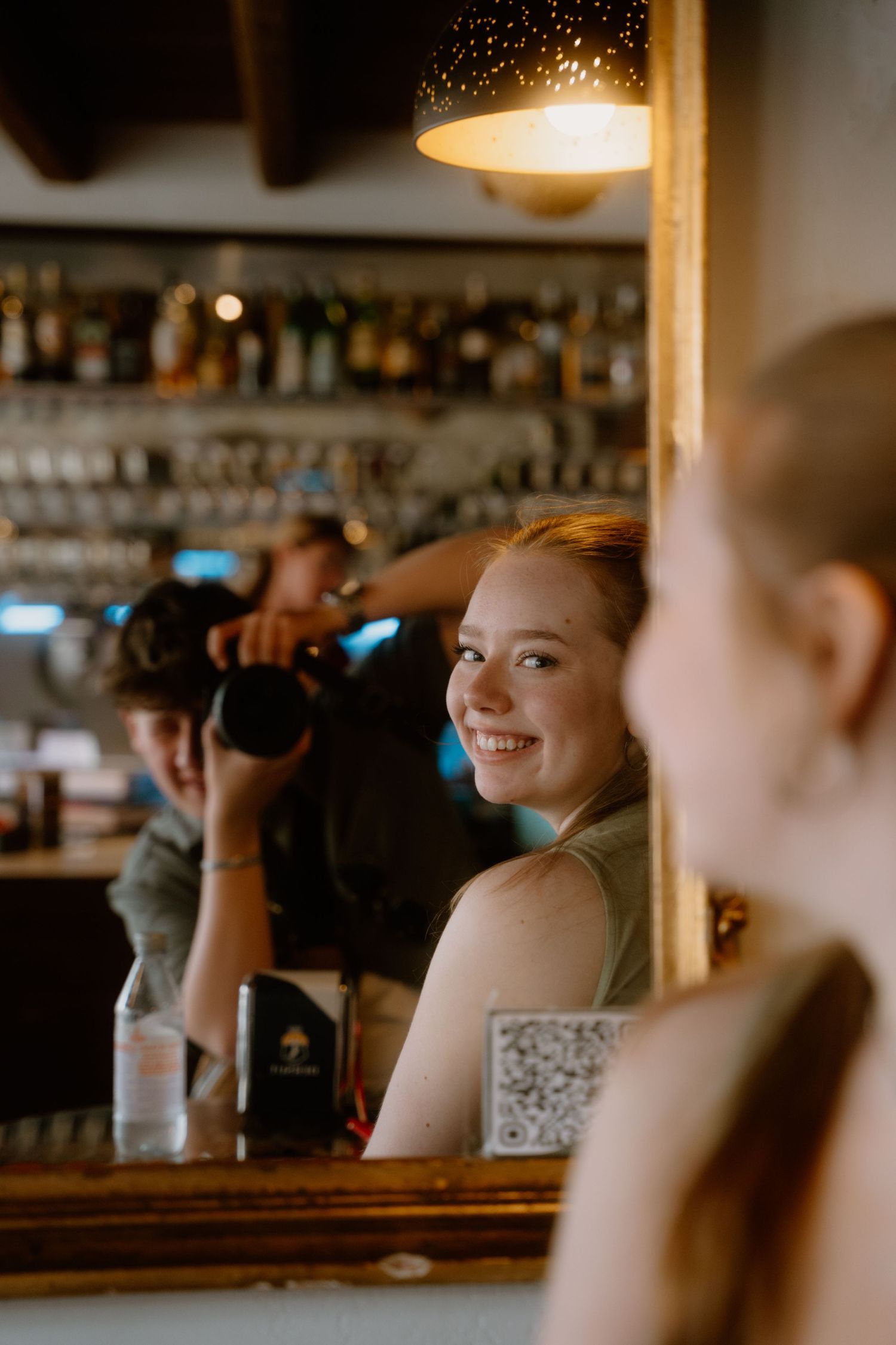 Inside a warmly lit bar, a camera captures someone smiling in a mirror reflection.