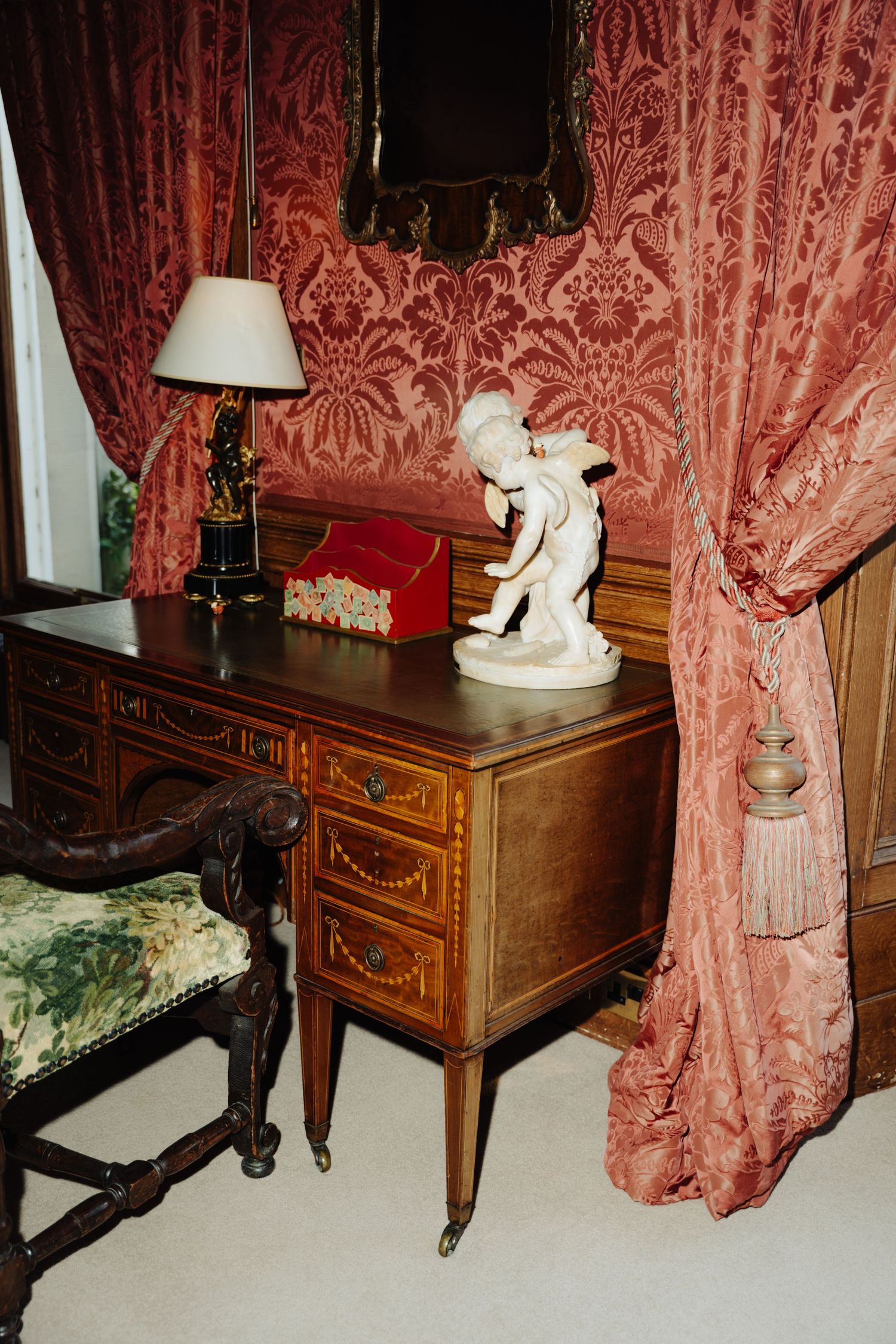 Elegant antique desk with red damask curtains, table lamp and decorative white statue in a Victorian style room.