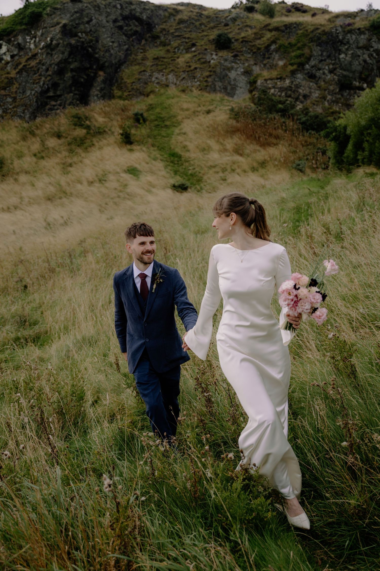 Bride in white gown holding pink bouquet walks hand-in-hand with groom in navy suit through grassy highland field.