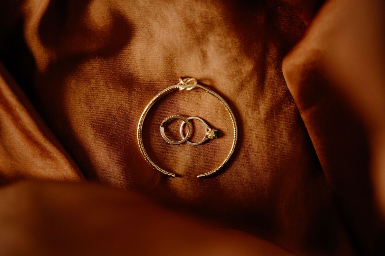 Two gold wedding rings nestled inside a circular bracelet on warm brown fabric.