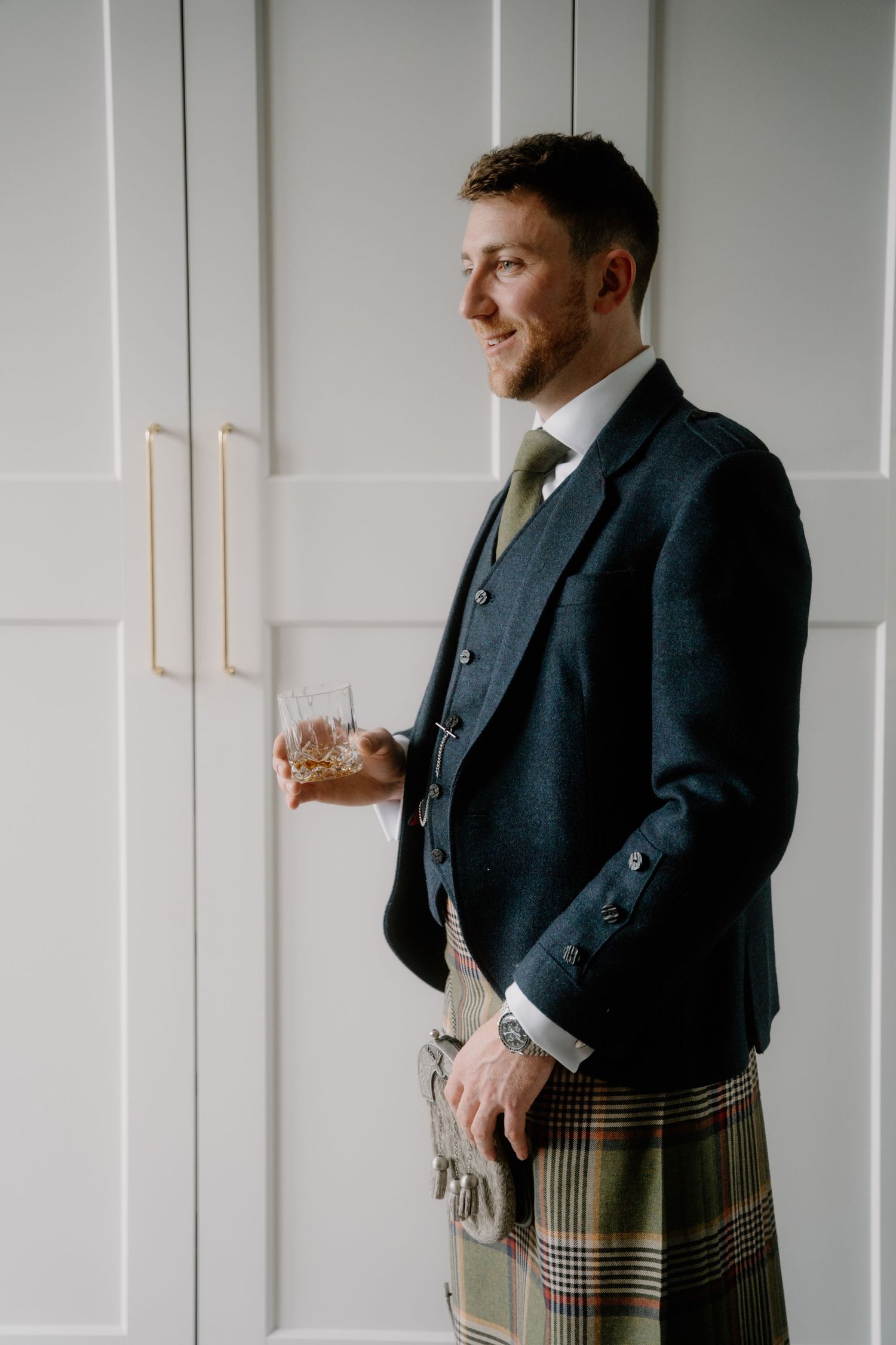 Person in traditional Scottish formal wear holding whiskey glass against white paneled wall.