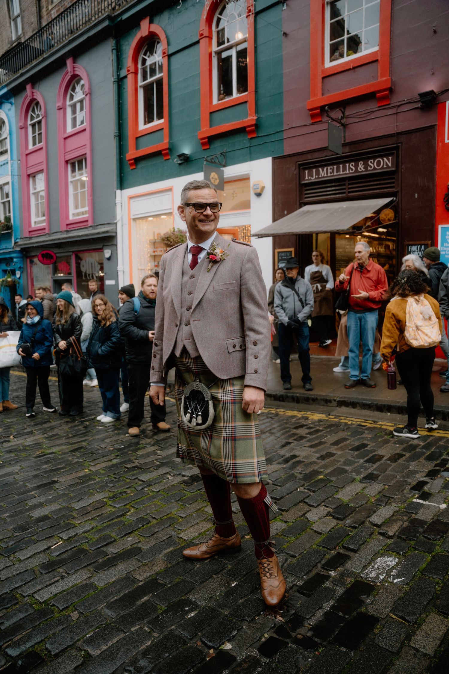 A person in traditional Scottish formal wear poses on a wet cobblestone street with colorful buildings behind.