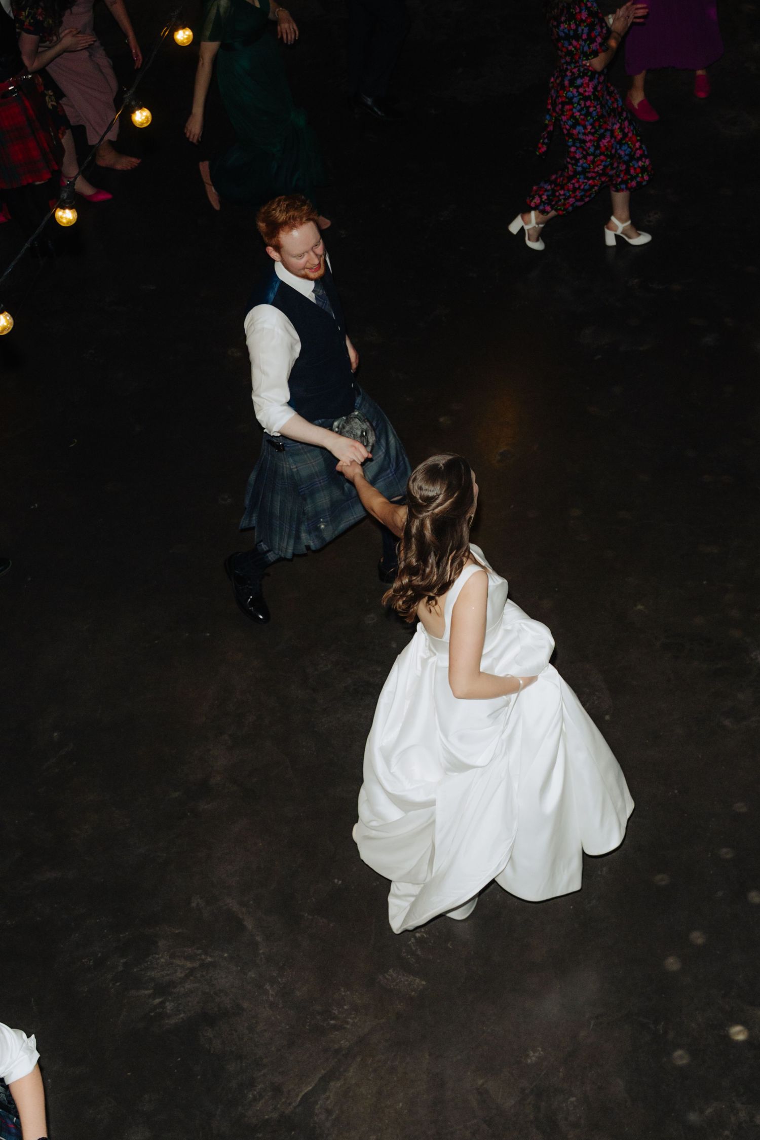 A couple dances together at a wedding reception, with the bride in a white gown and partner wearing traditional Scottish attire.