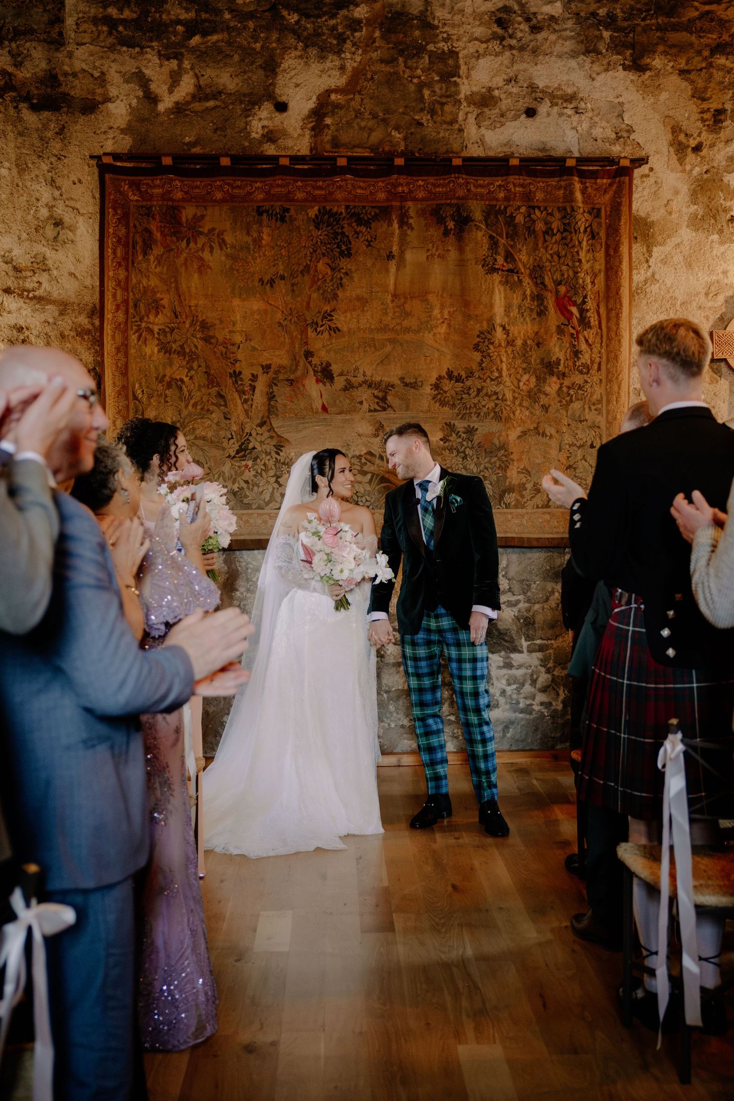 Bride and groom walk down the aisle in a rustic stone castle venue, guests applauding, groom in tartan trousers.