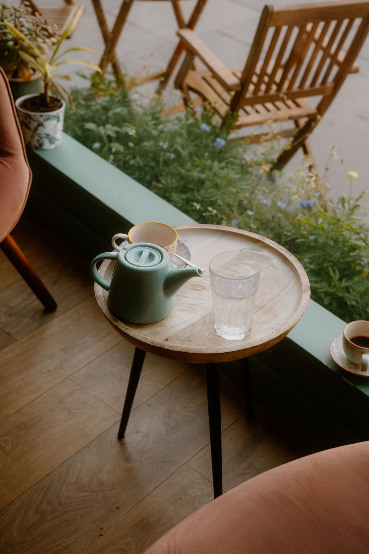 A cozy coffee setup with a green teapot on a round wooden table near garden chairs.