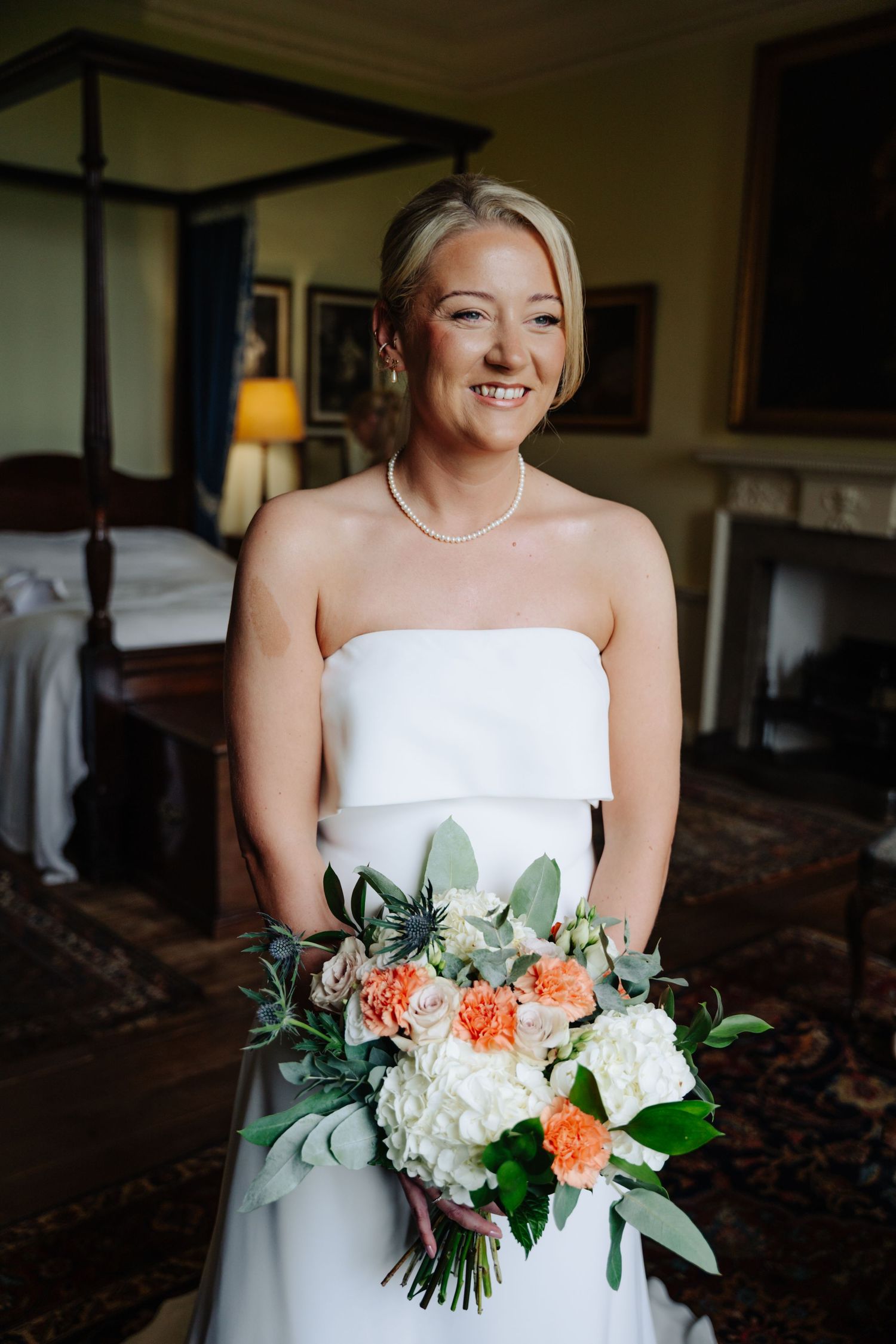 Bride in strapless white wedding dress holds a beautiful bouquet with coral and white flowers against dark interior. Boswell Estate