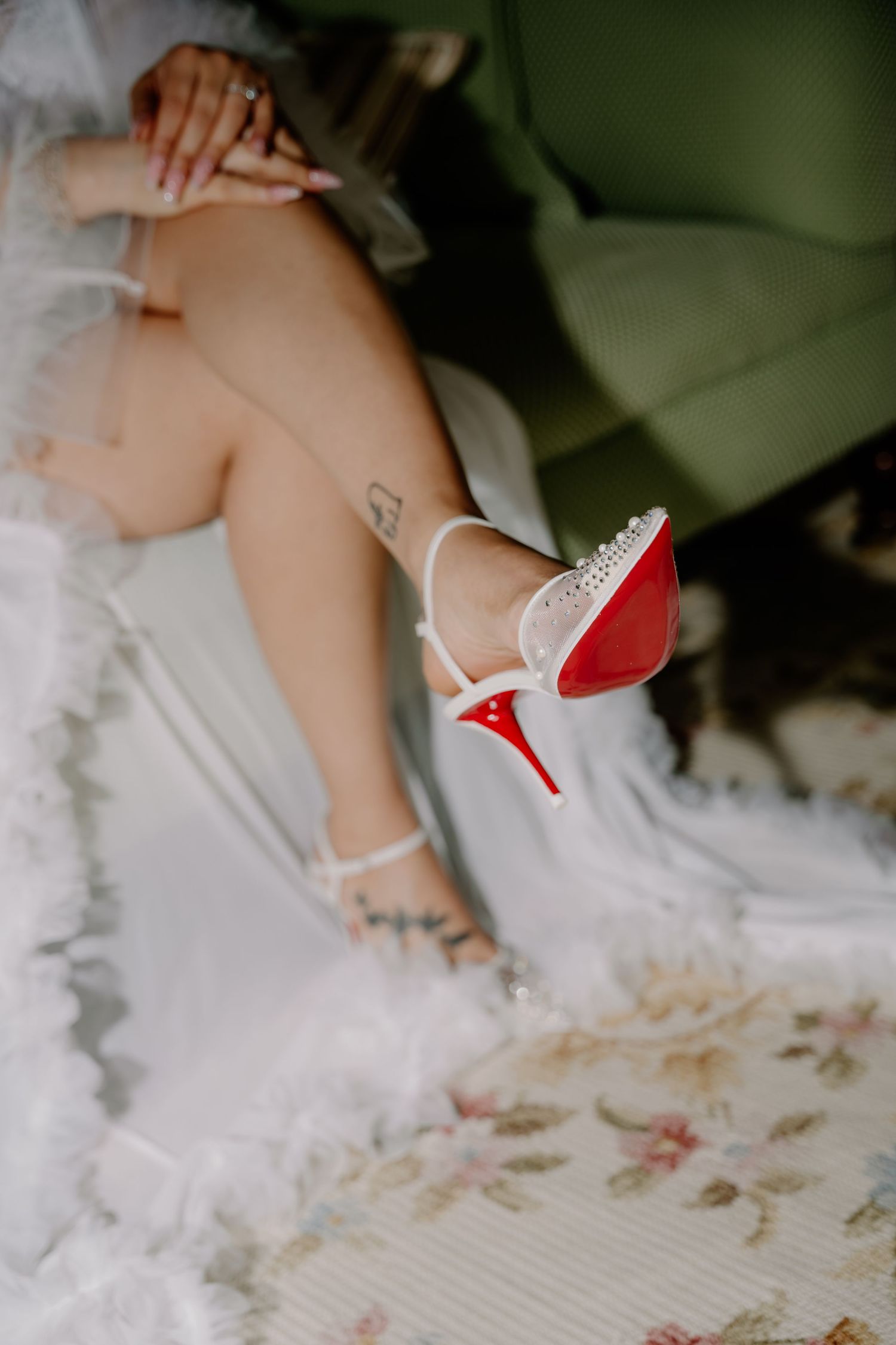 Bride wearing white Christian Louboutin heels with red soles and pearl details, sitting in green chair with wedding dress.