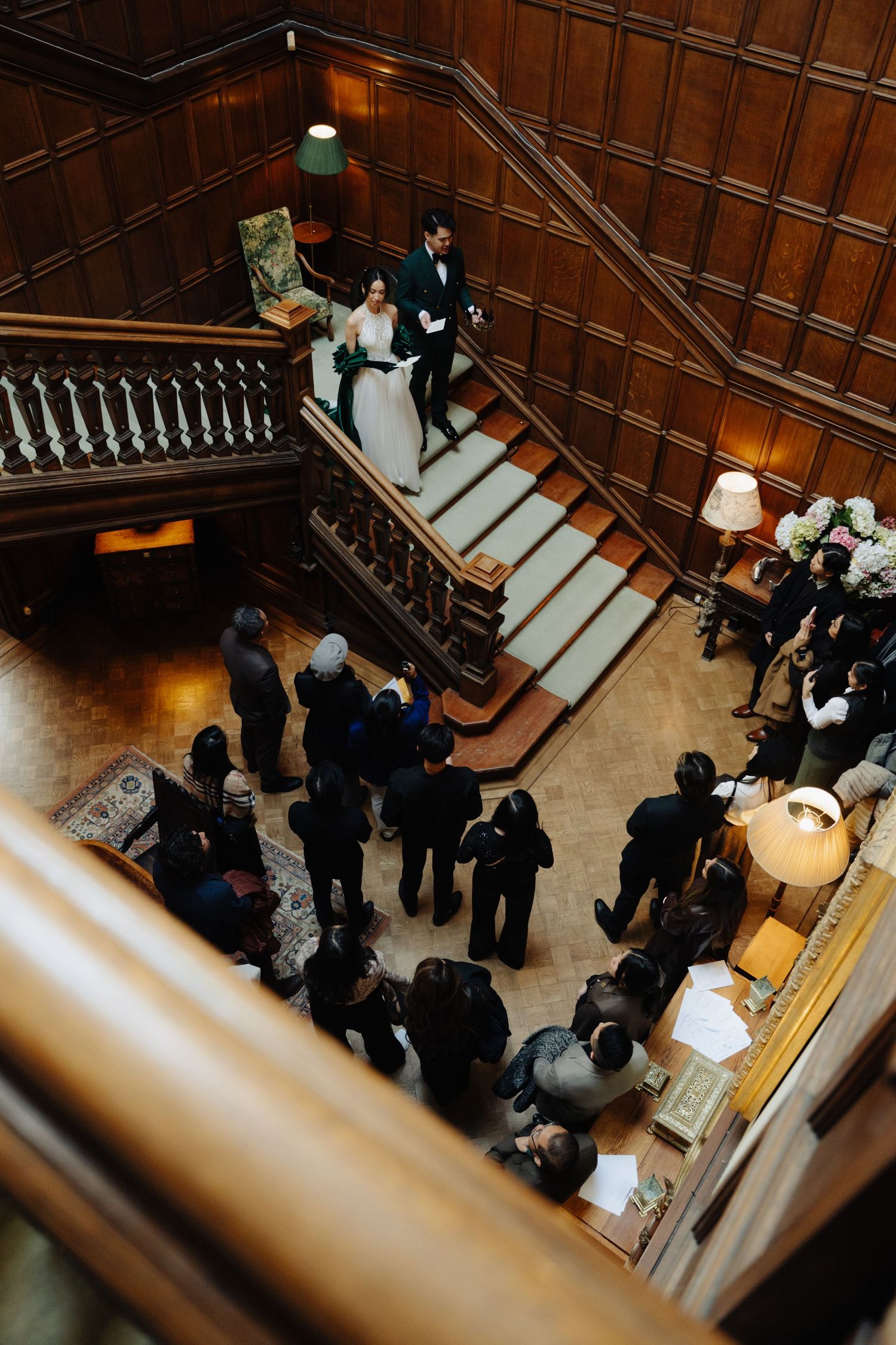 Aerial view of bride and groom descending grand wooden staircase surrounded by wedding guests in elegant venue.