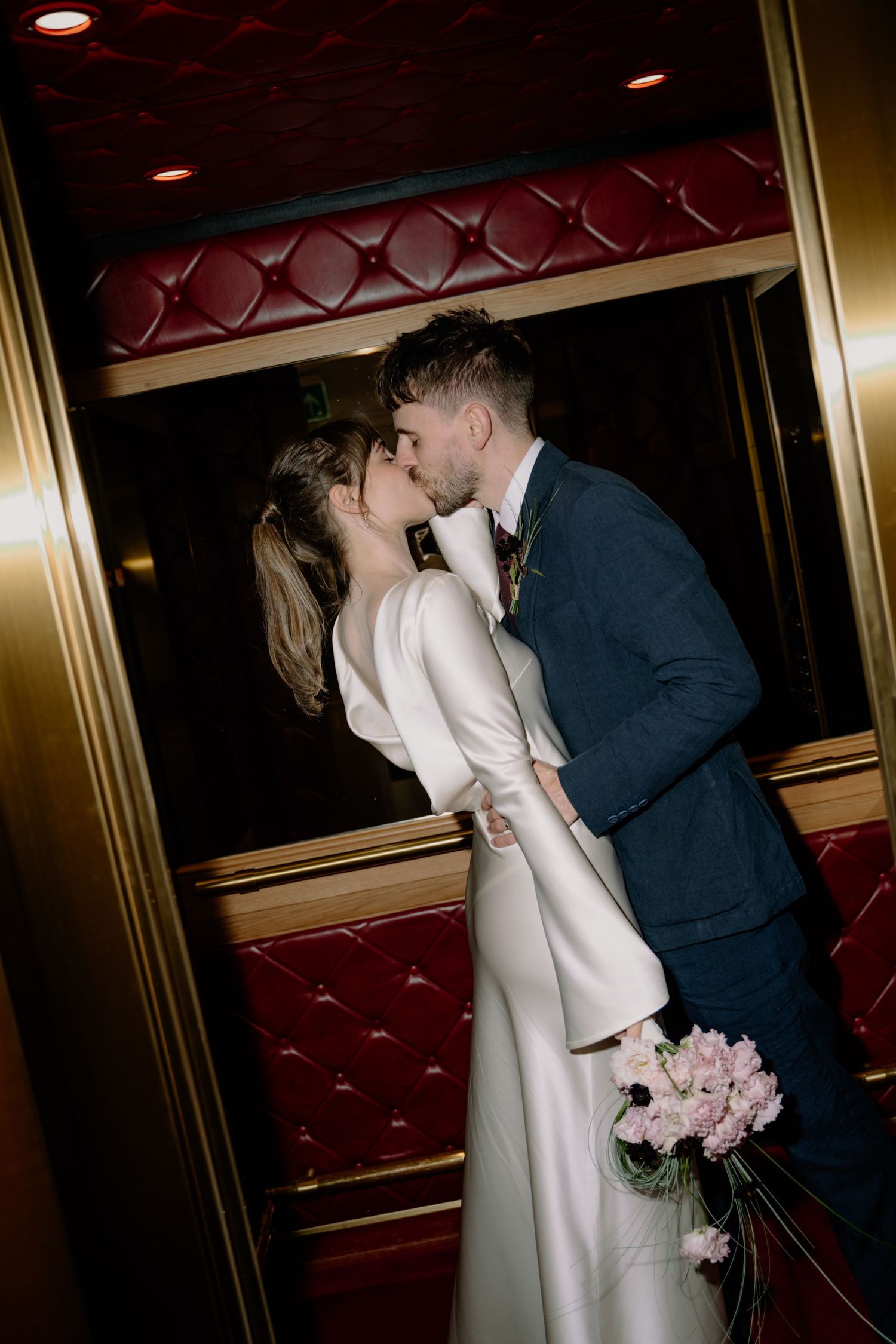 Bride and groom share a romantic kiss in an elegant gold and red elevator, bride holding pink peonies bouquet.
