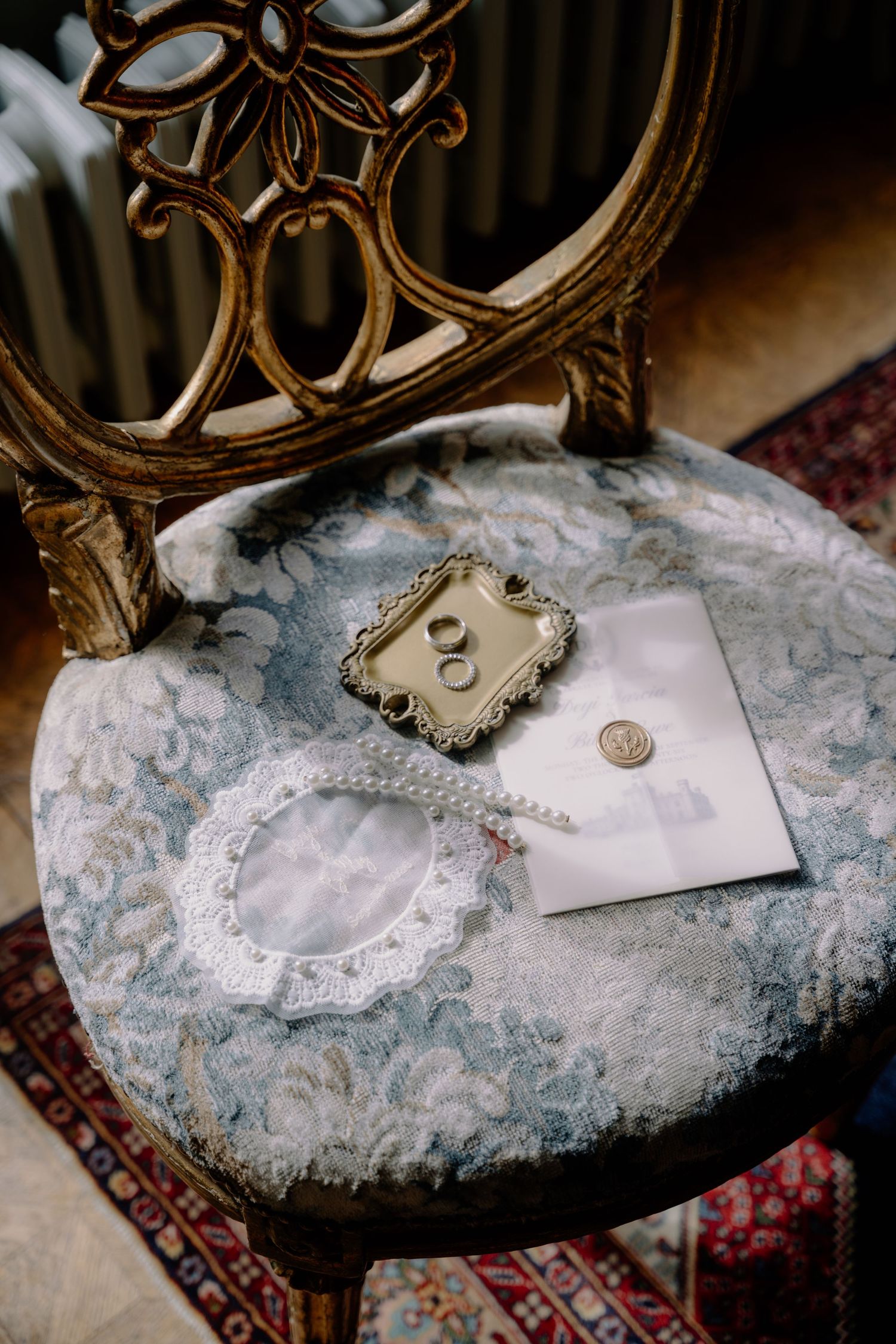 Vintage ornate wooden chair with wedding rings on gold tray, wax-sealed envelope, and lace garter on blue velvet seat.