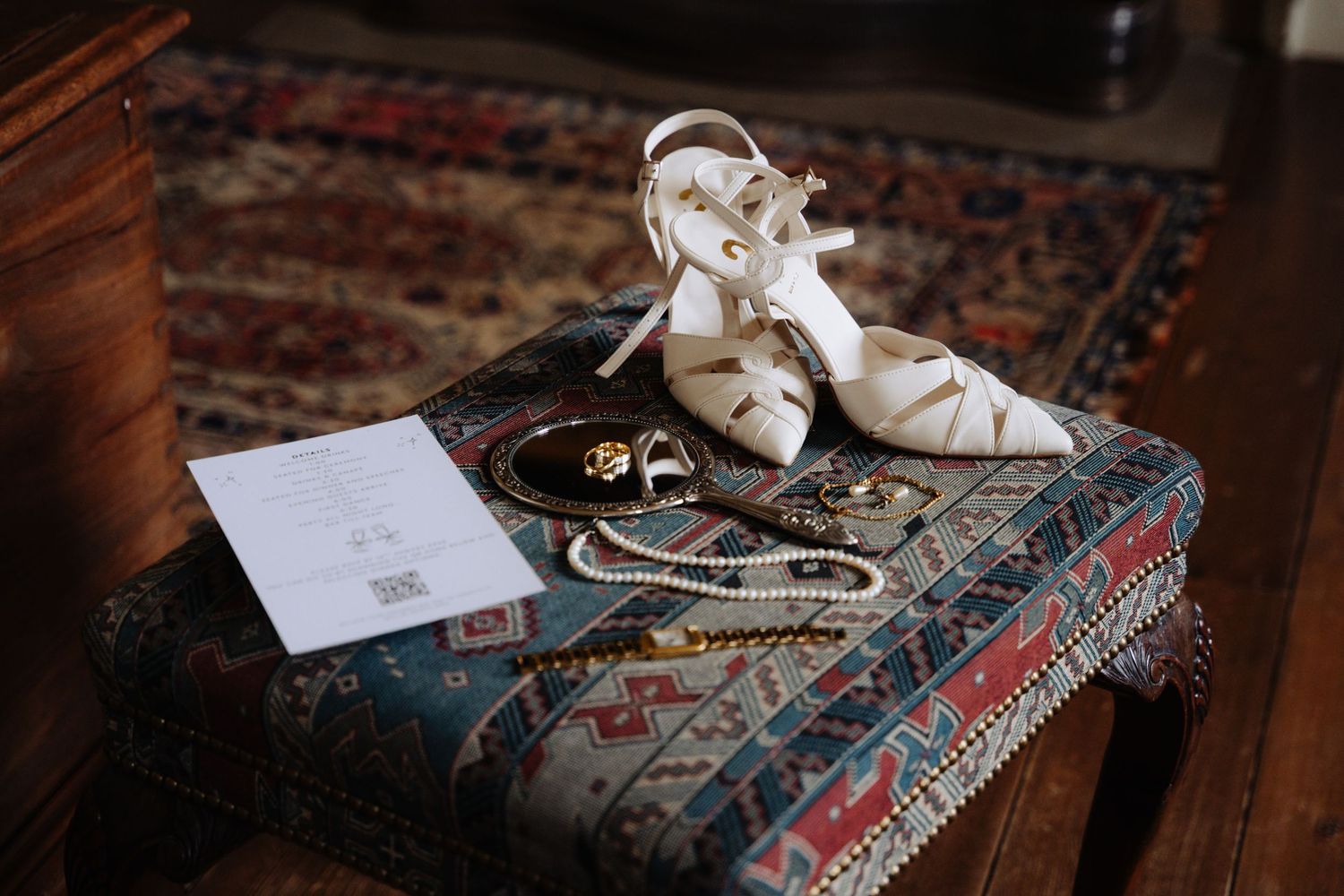White wedding shoes and jewelry arranged on a decorative patterned rug with an invitation card.