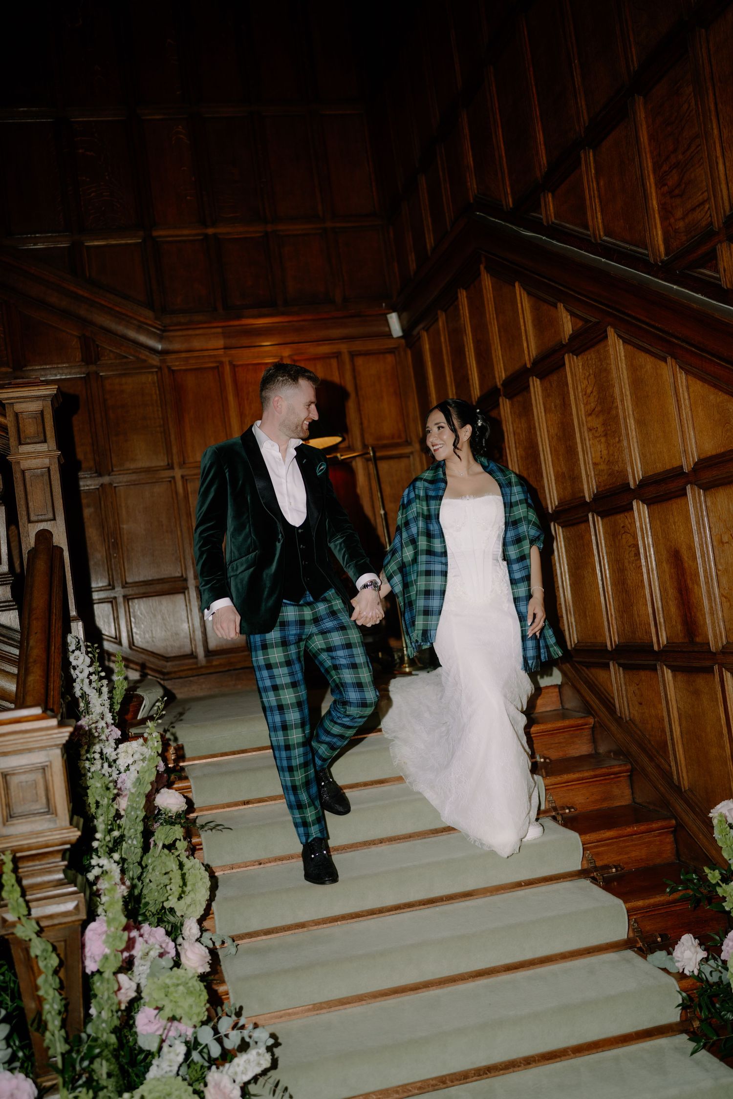 A couple in Scottish tartan attire descends a grand wooden staircase, holding hands and smiling on their wedding day.