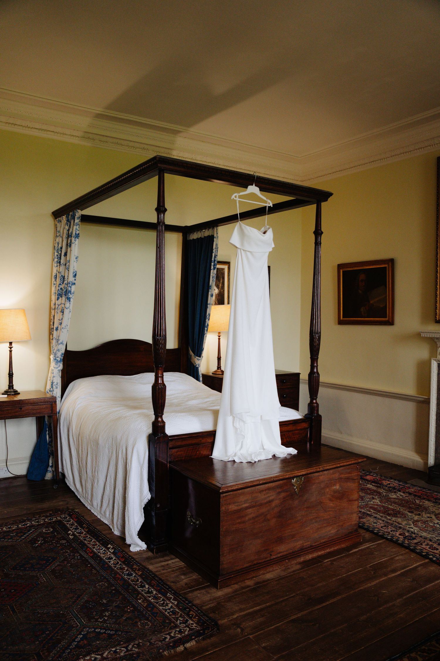 A white wedding dress hangs elegantly beside a four-poster bed in a traditional bedroom with dark hardwood floors. Boswell Estate