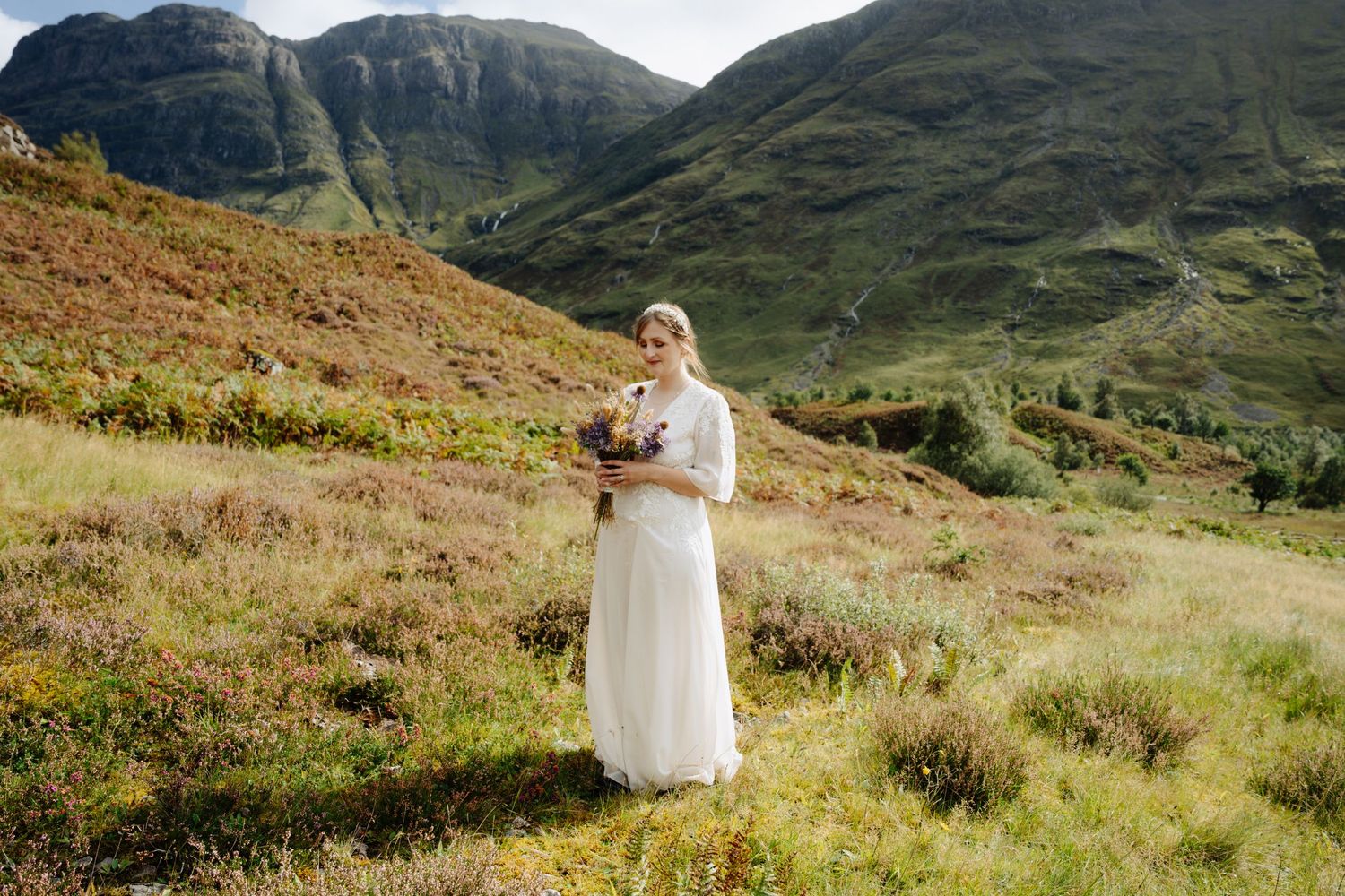 A figure in a flowing white dress stands in a scenic Scottish highland landscape with mountains in the background.