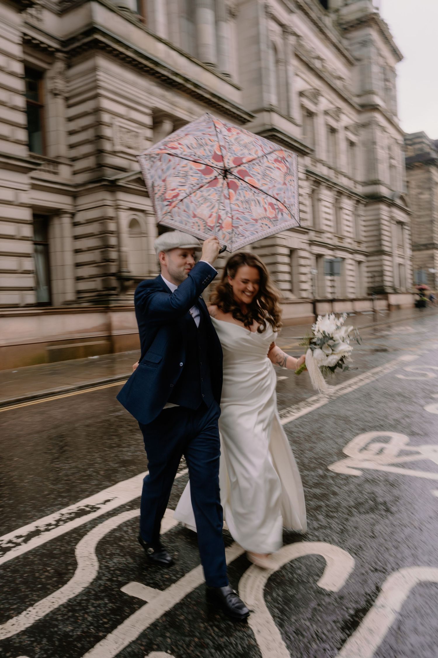 Bride and groom crossing a rainy street holding a floral umbrella, bride in white gown with bouquet, groom in navy suit.